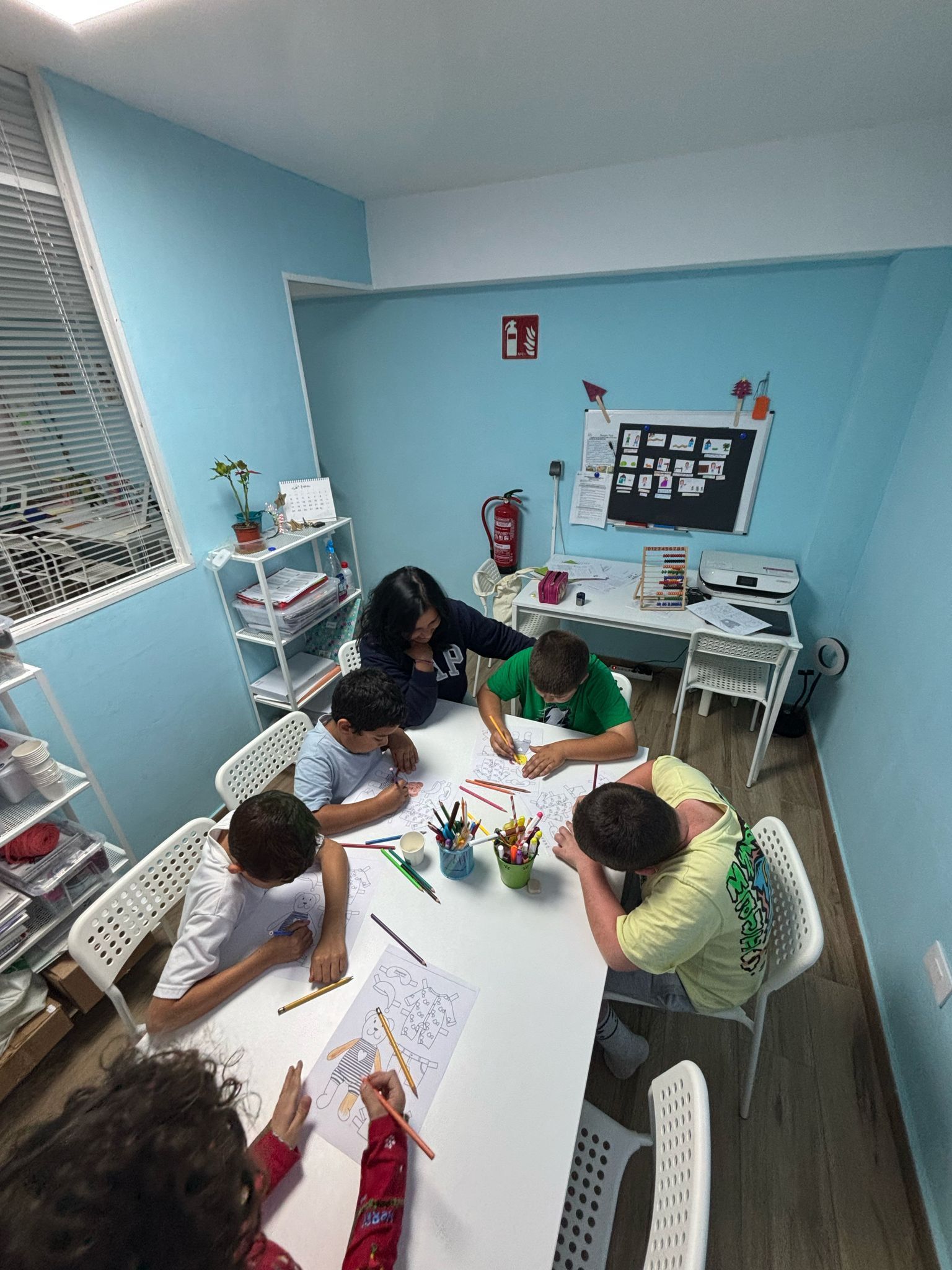 Niños y maestra alrededor de una mesa dibujando en una habitación de color azul claro.