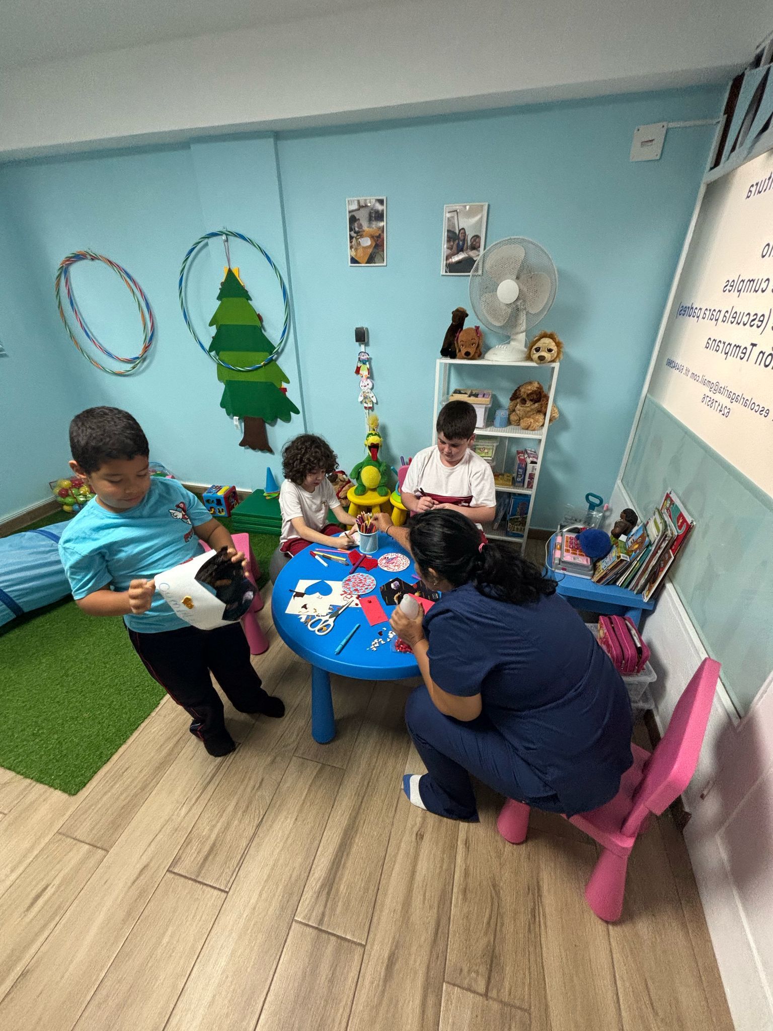 Niños y un adulto jugando en una mesa. Un niño sostiene un juguete. La habitación está decorada y hay un ventilador.