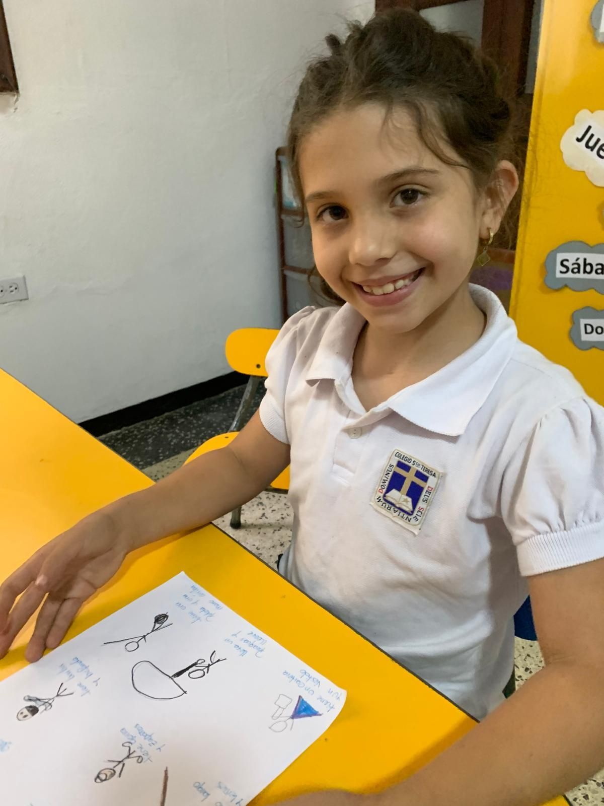 Una joven de cabello castaño sonríe, sentada en un escritorio amarillo, sosteniendo un dibujo y vistiendo un uniforme blanco.