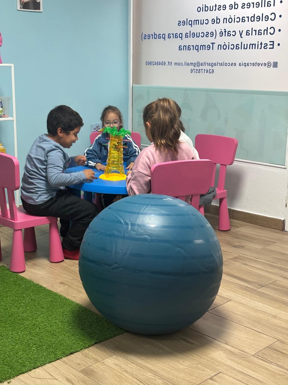 Niños jugando en una mesa en una habitación con paredes azules. Hay sillas rosas y una pelota de ejercicio azul.