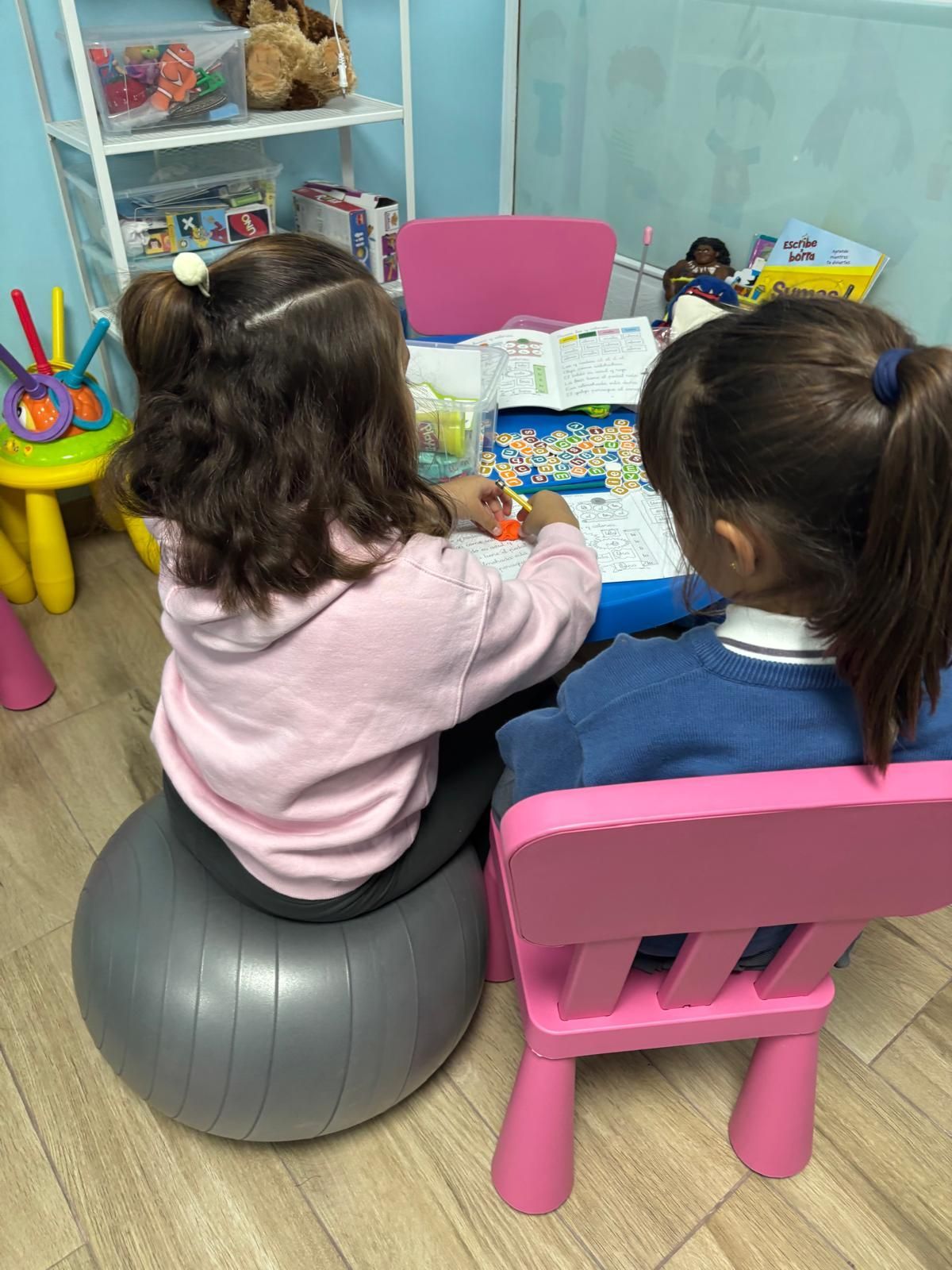 Dos chicas jóvenes en una mesa azul, una en una pelota de ejercicio gris y otra en una silla rosa, trabajando juntas.