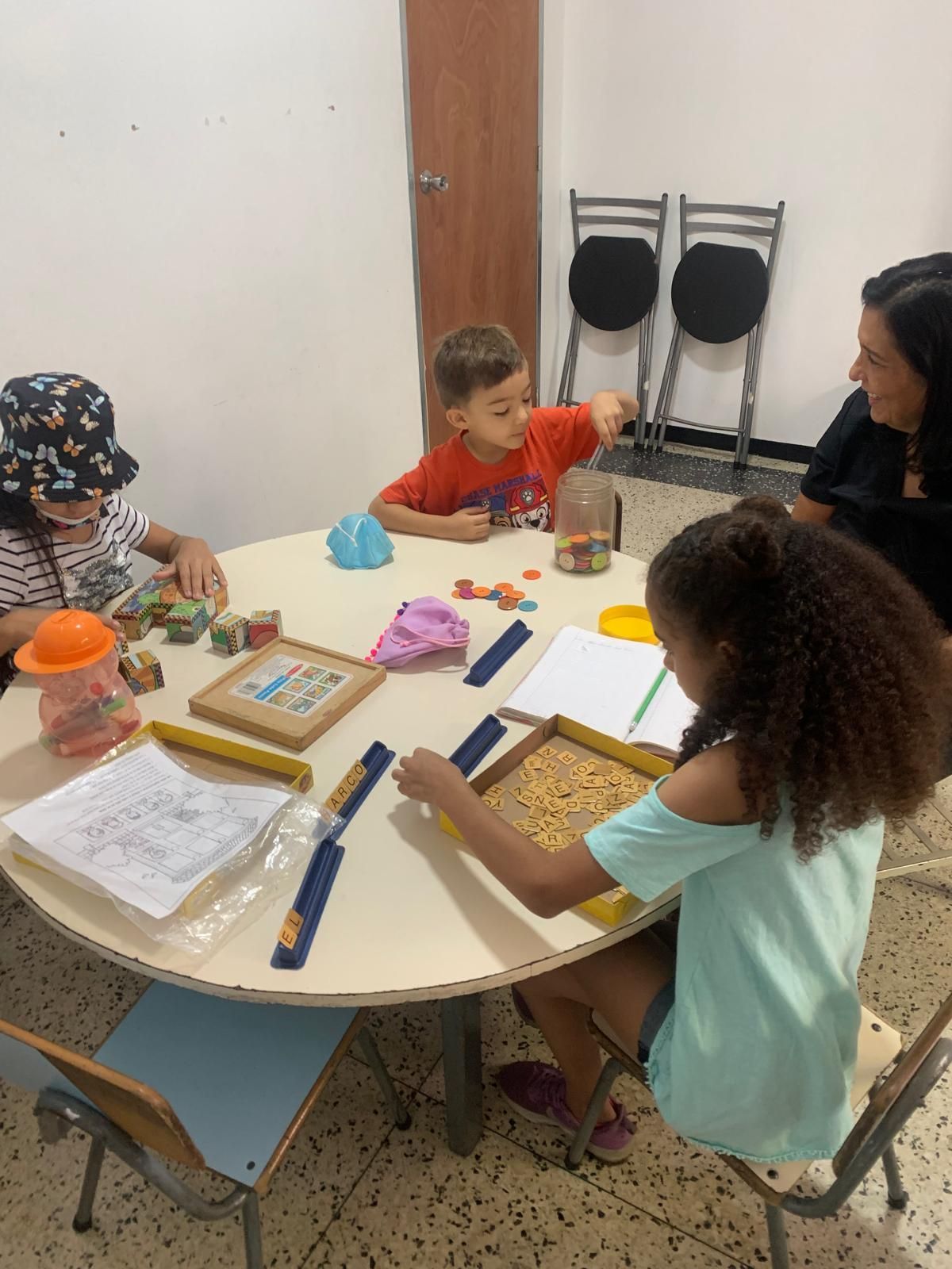 Niños en una mesa redonda con un adulto, jugando con juguetes y escribiendo en cuadernos.