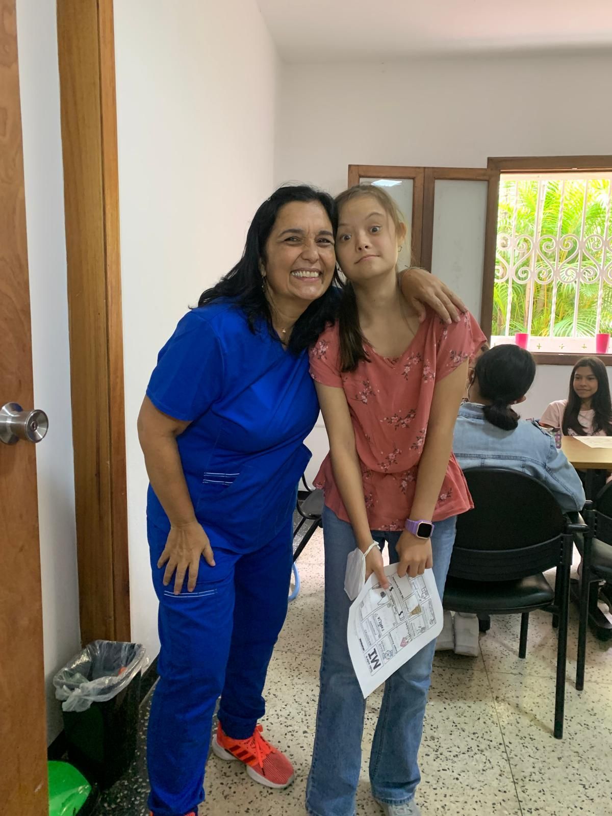Una mujer con uniforme azul abraza a una niña; ambas sonríen. Un aula con otros niños al fondo.