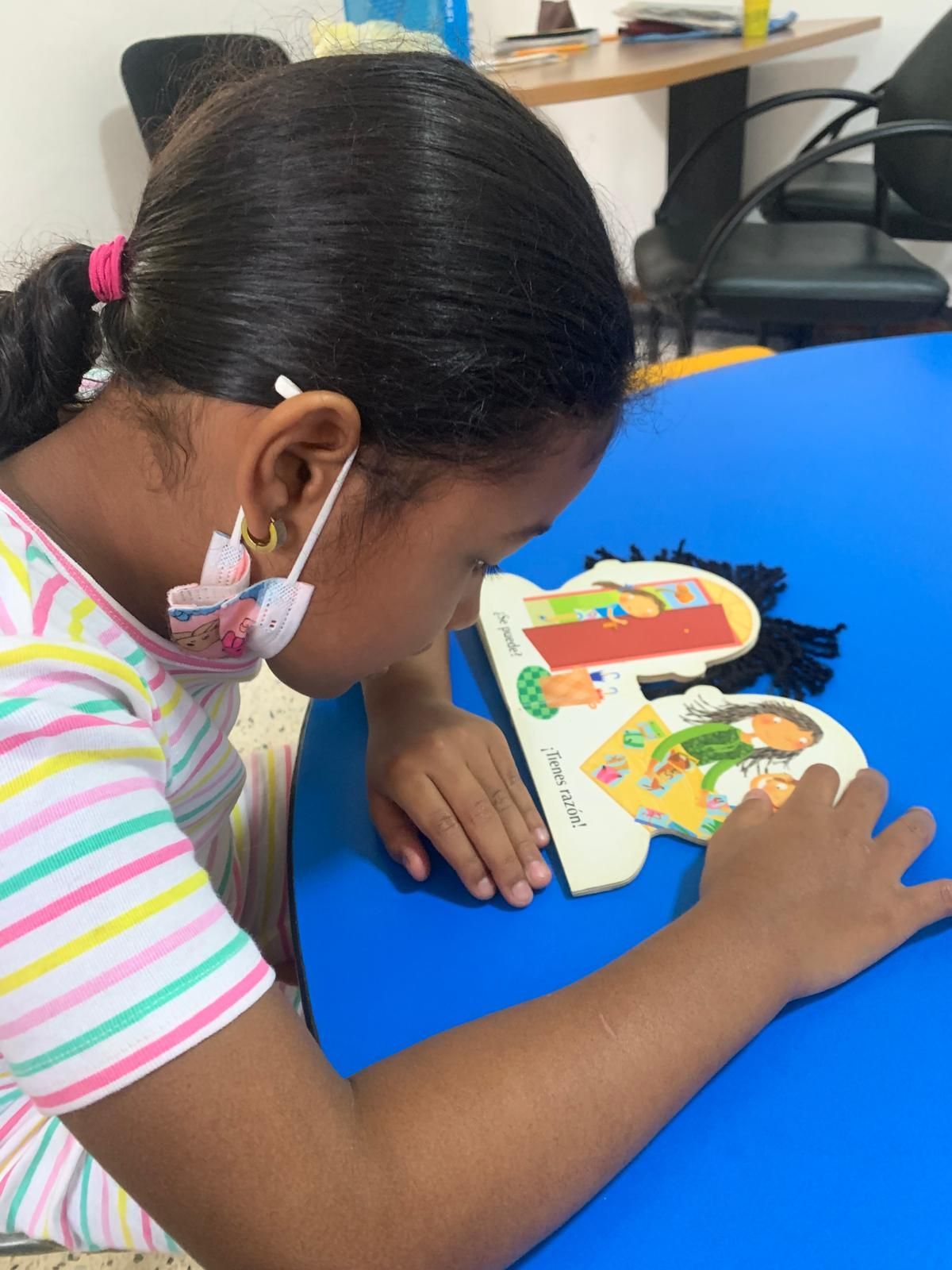 Chica con cabello oscuro y máscara, se inclina sobre un rompecabezas colorido sobre una mesa azul.