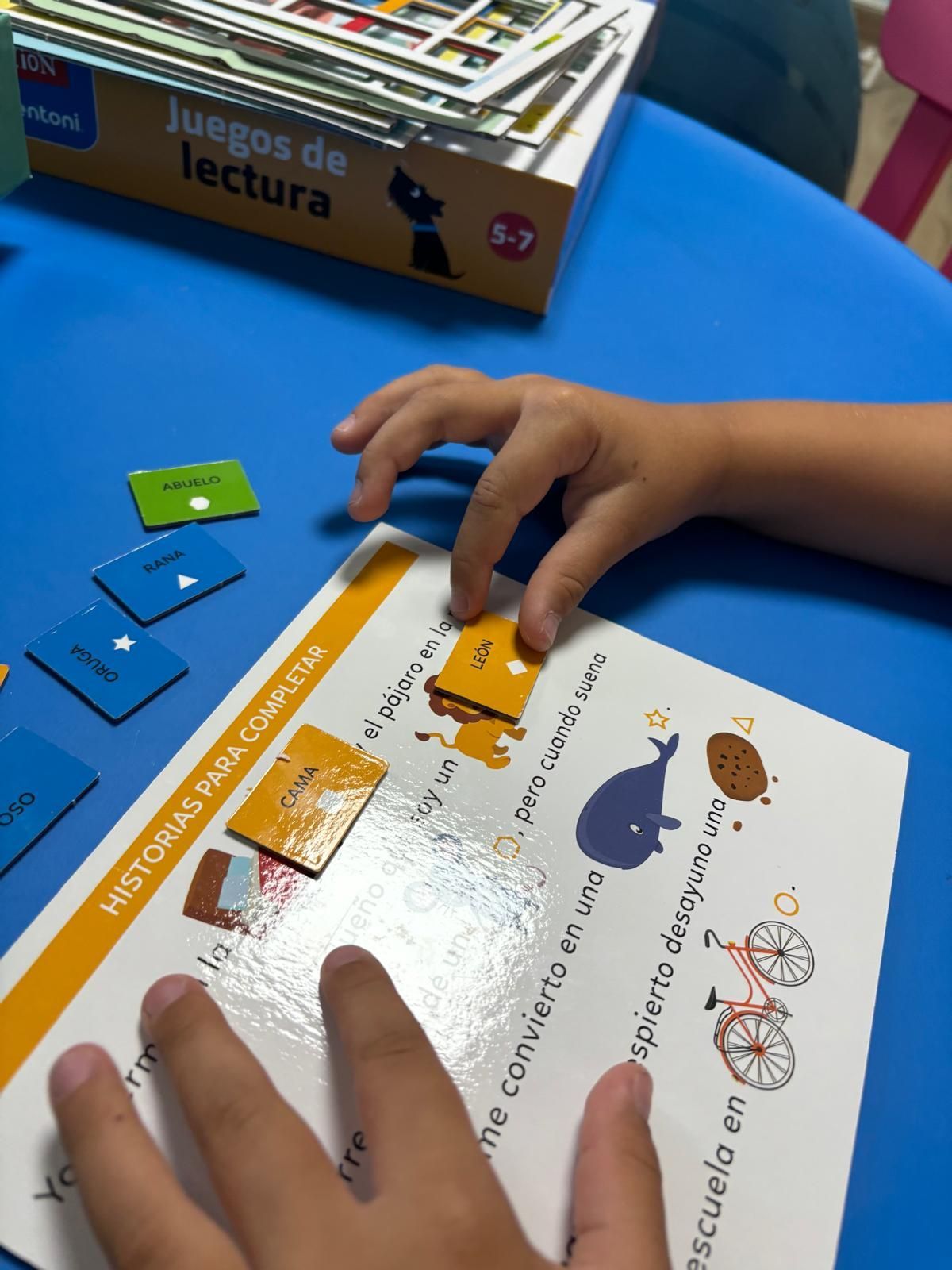 Un niño juega a la lectura, colocando fichas en una página. El ambiente es interior, sobre una mesa azul.