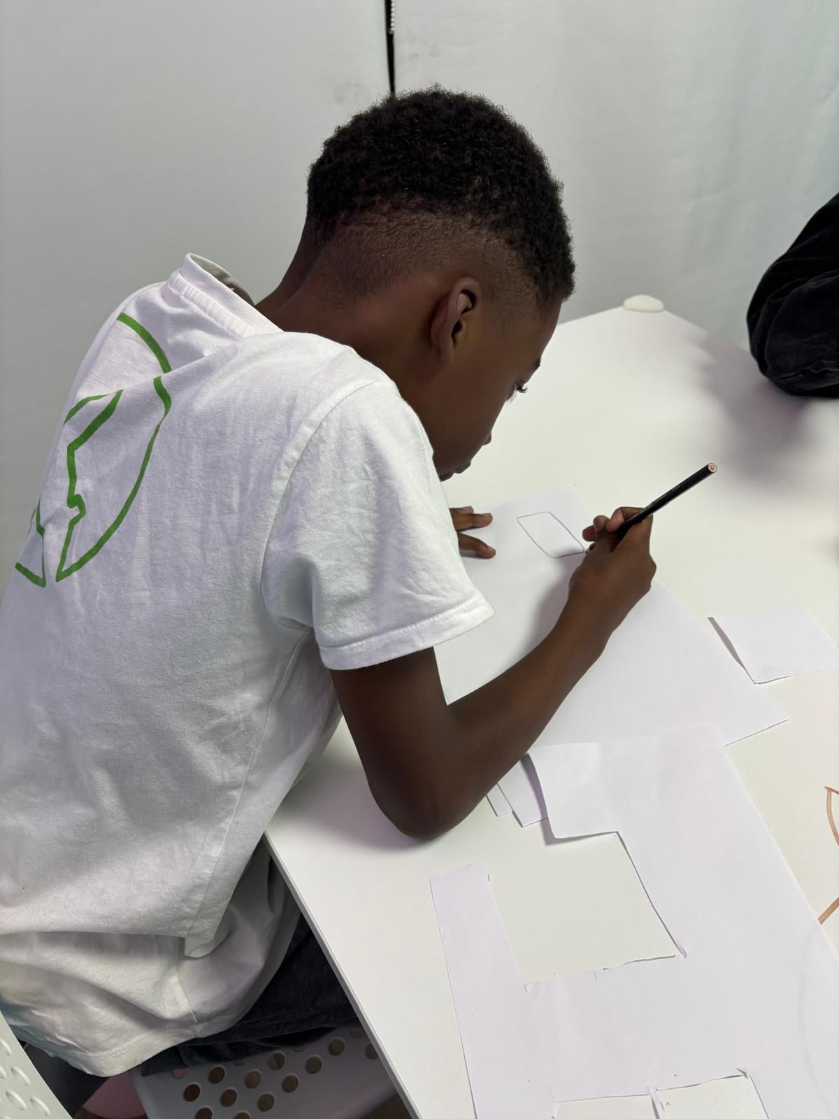 Un joven negro con una camisa blanca dibuja con un lápiz sobre papel en una mesa blanca.