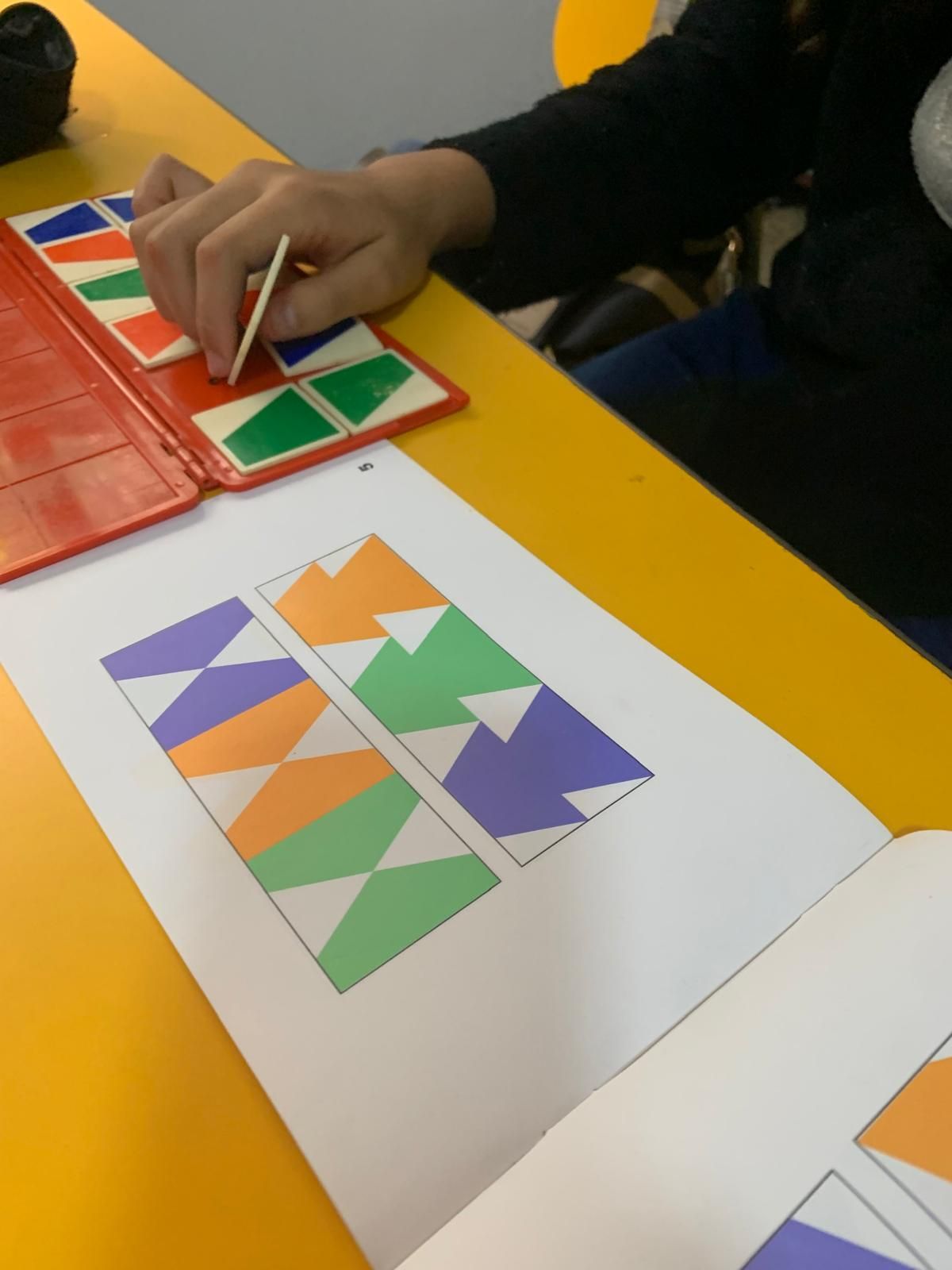 Persona que utiliza piezas de rompecabezas de colores para hacer coincidir un diagrama en un papel, sobre una mesa amarilla.