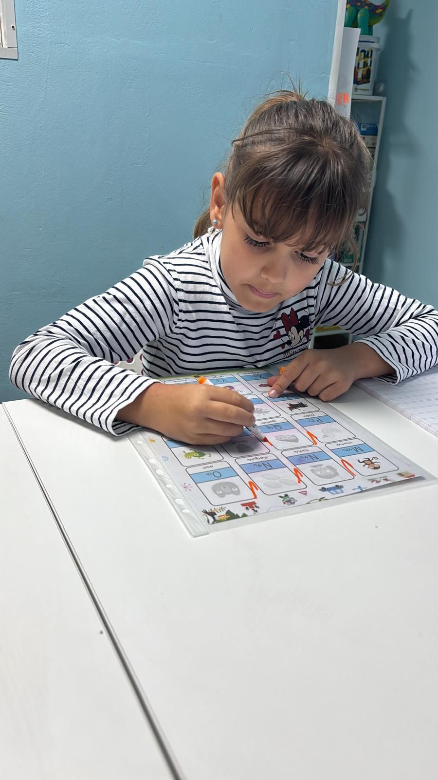 Chica con cabello castaño con camisa a rayas, escribiendo en una hoja de trabajo en una mesa blanca.