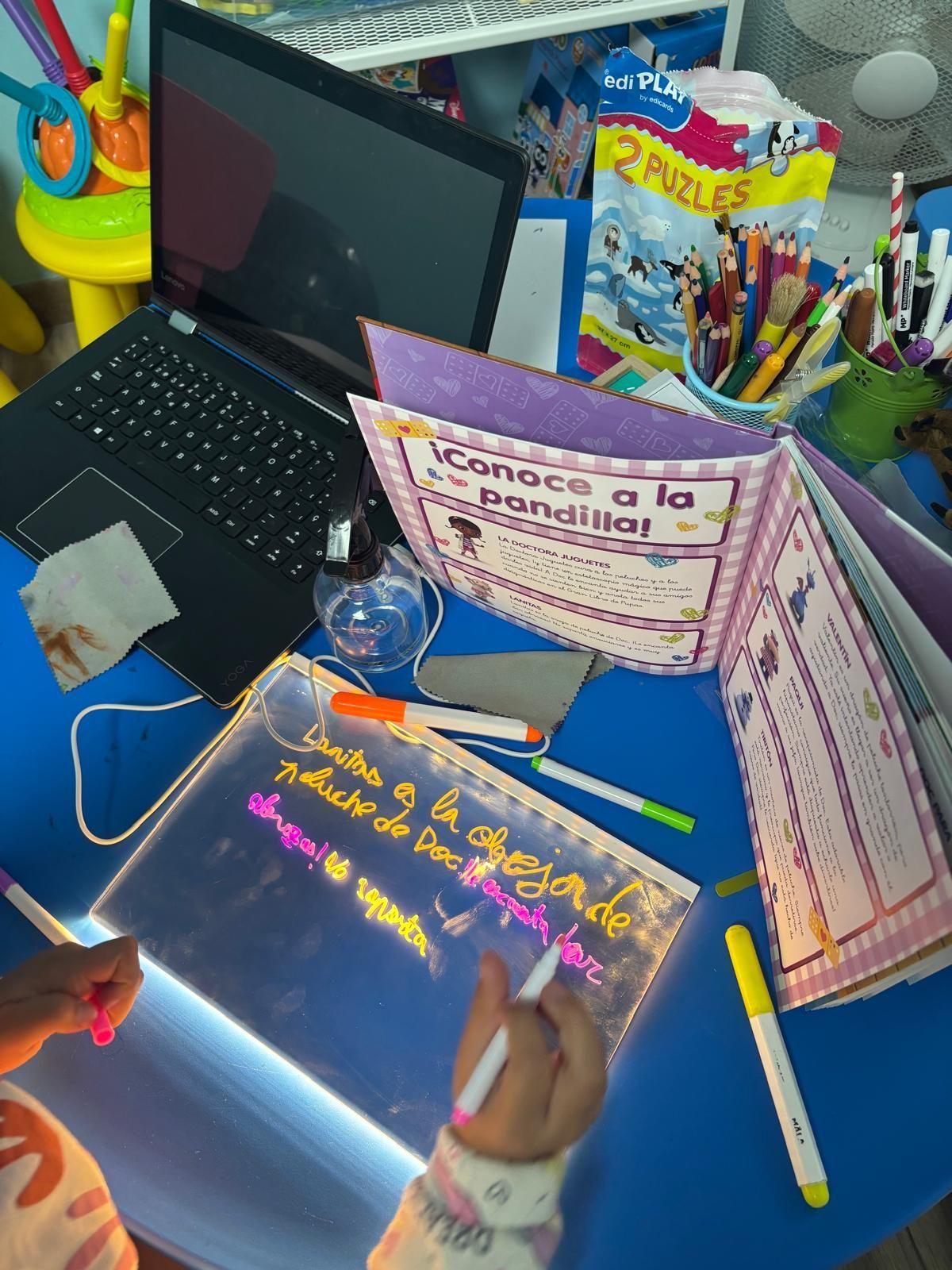 Niño escribiendo en una pizarra luminosa en un escritorio con una computadora portátil, libros y marcadores.