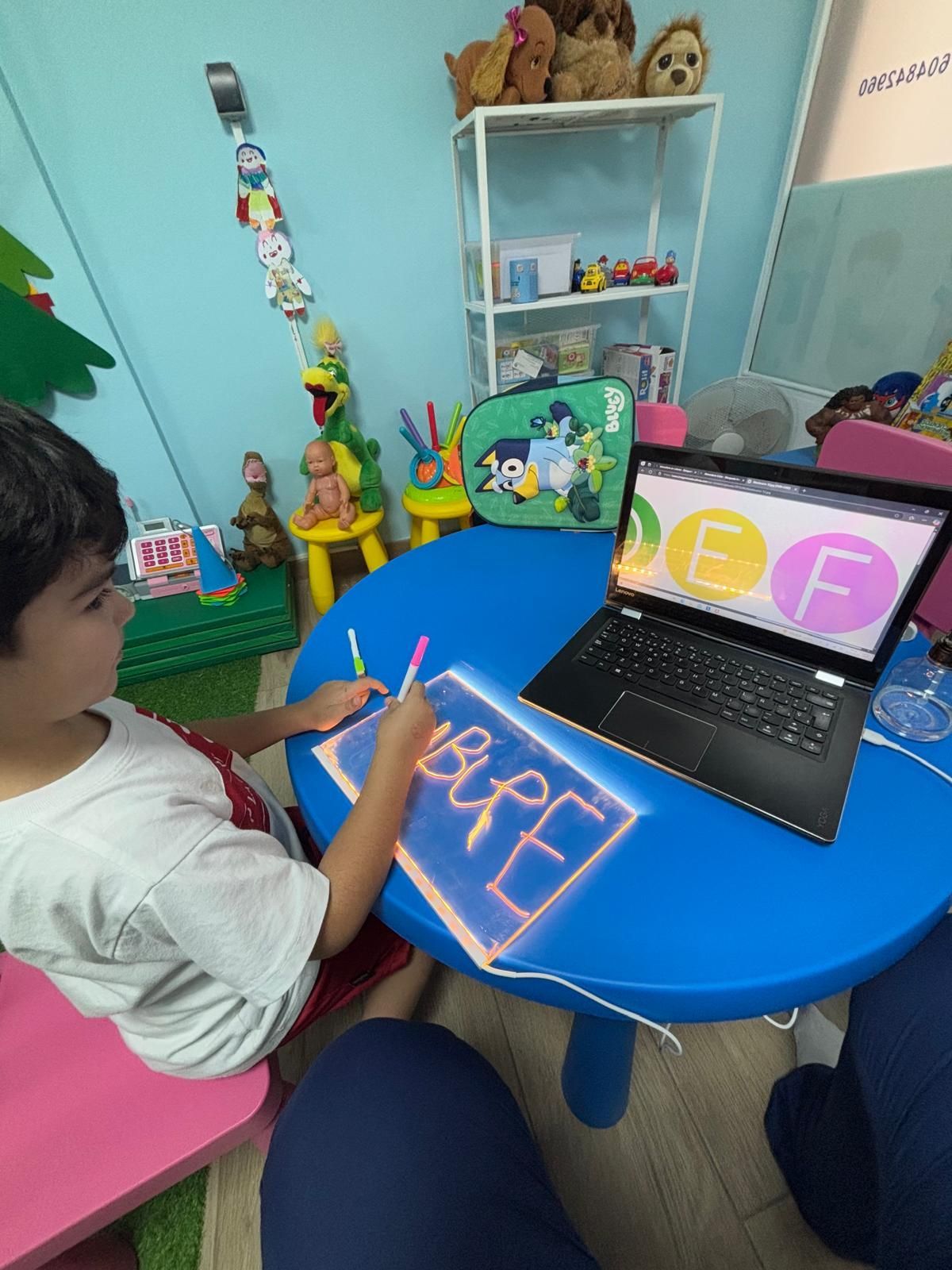 Un niño pequeño aprendiendo letras en una mesa azul con una computadora portátil, dibujando con un bolígrafo de neón.