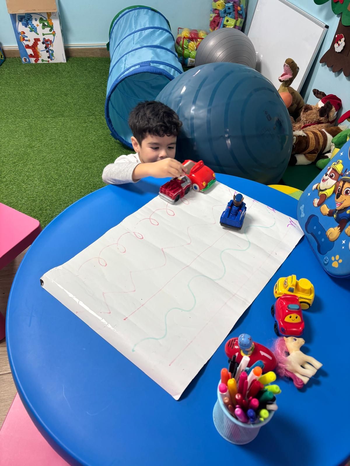 Un niño pequeño juega con coches de juguete sobre una mesa azul, con marcadores y papel en una habitación colorida.