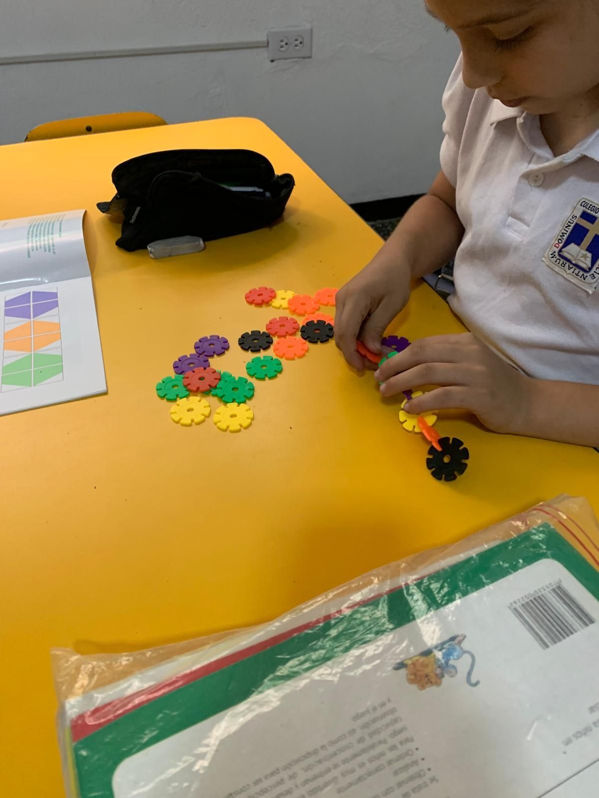 Niño usando formas entrelazadas de colores sobre una mesa amarilla en un salón de clases.