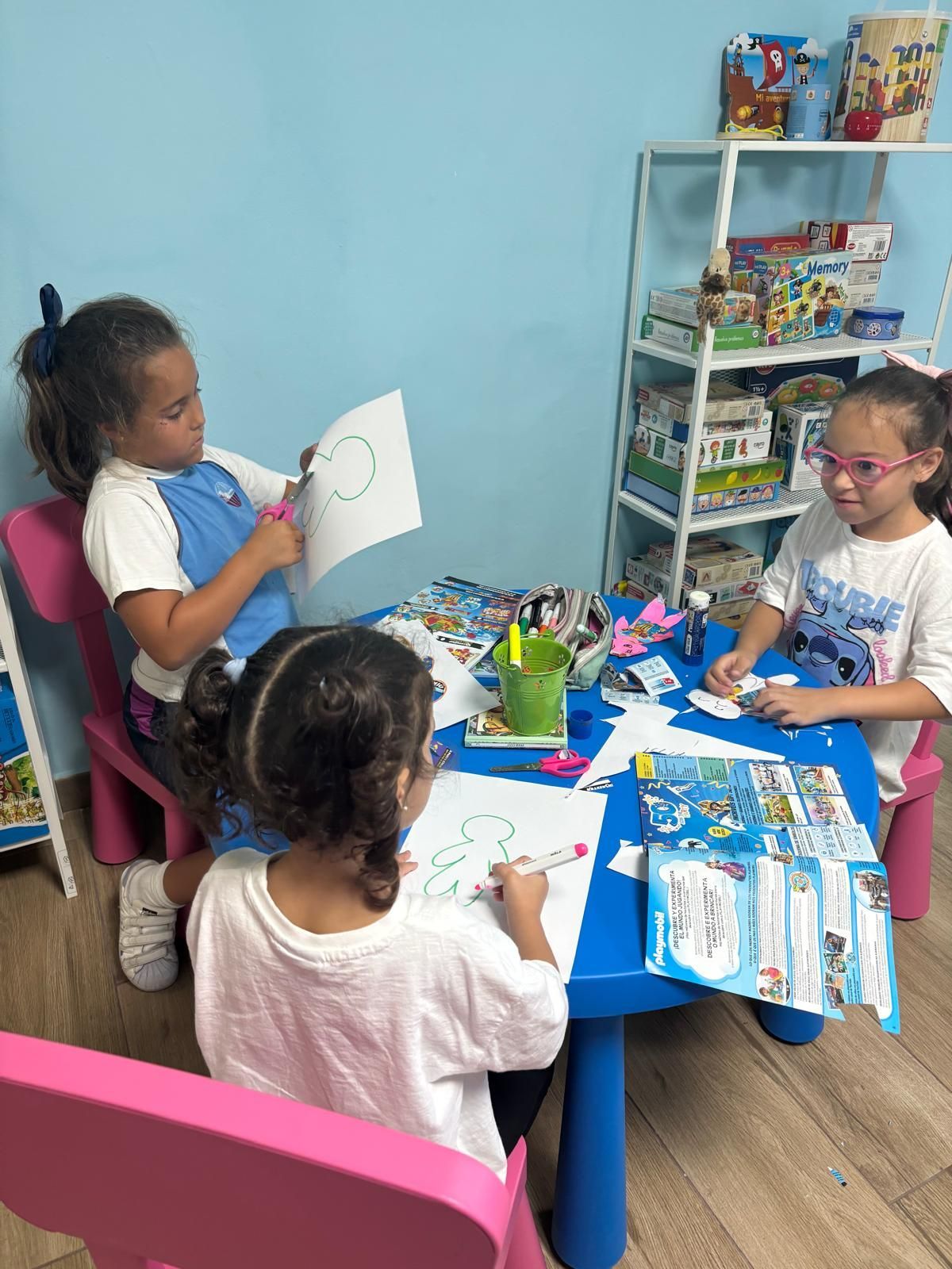 Tres niñas haciendo manualidades en una mesa azul, una cortando un círculo, la otra dibujando.