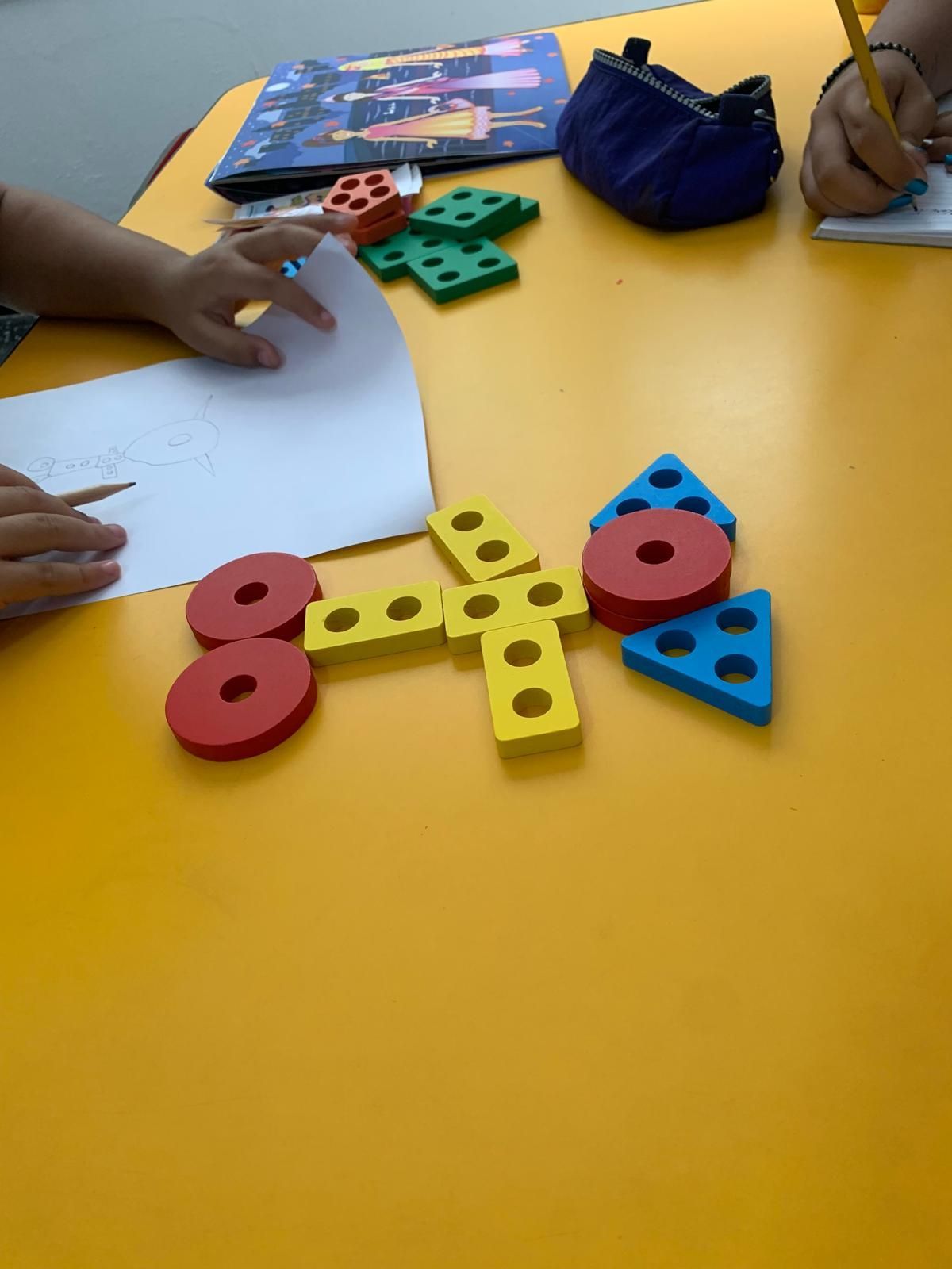 Manos con lápices dibujando sobre papel, formas geométricas de colores sobre una mesa amarilla.