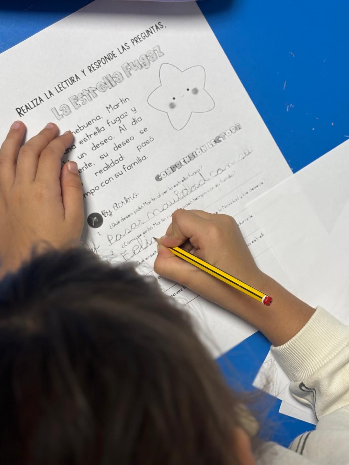 Manos de un niño escribiendo en una hoja de trabajo con un dibujo de estrella. Escritorio azul.