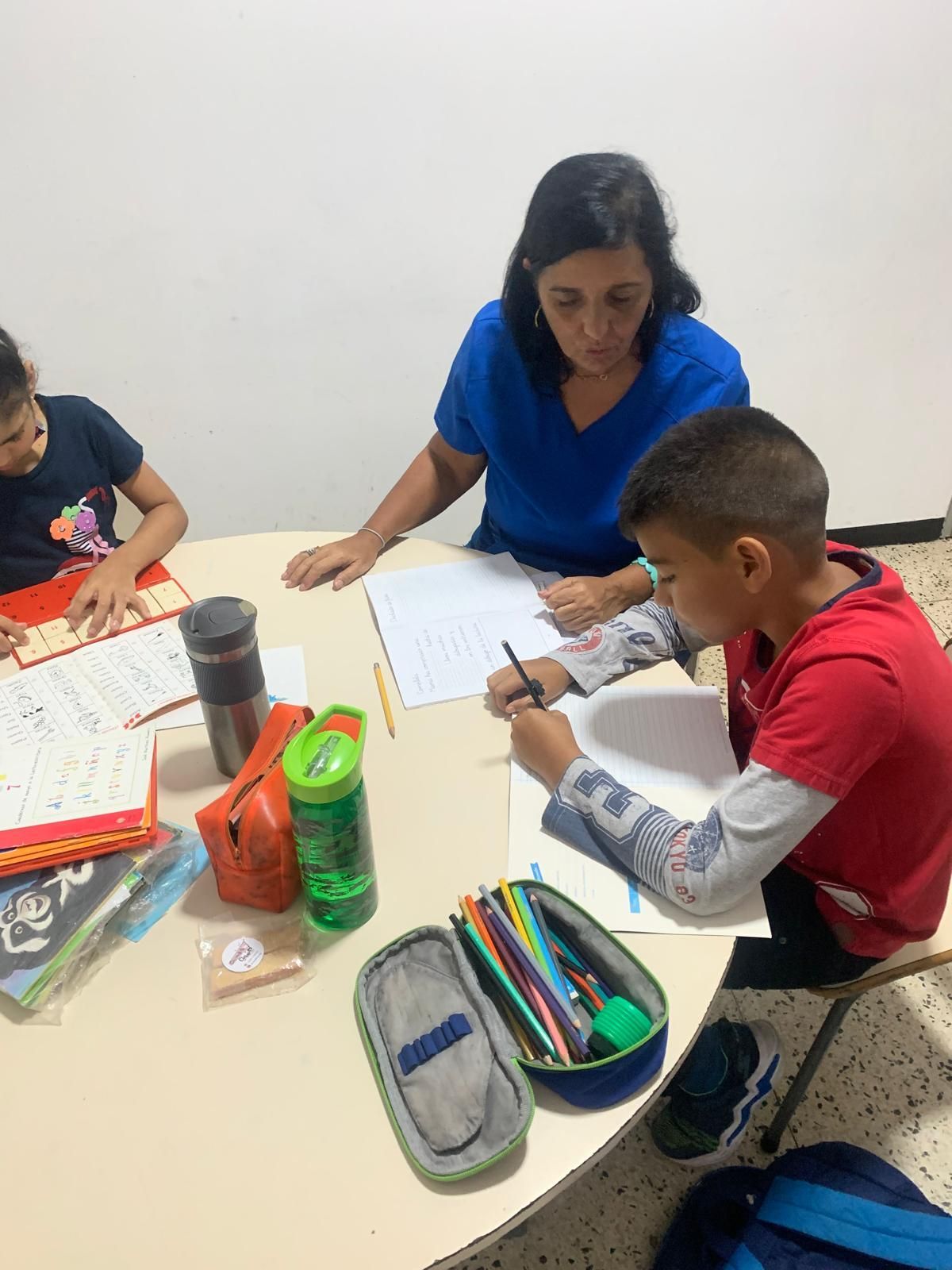 Una mujer da clases particulares a dos niños en una mesa. Uno dibuja, el otro trabaja en un libro.