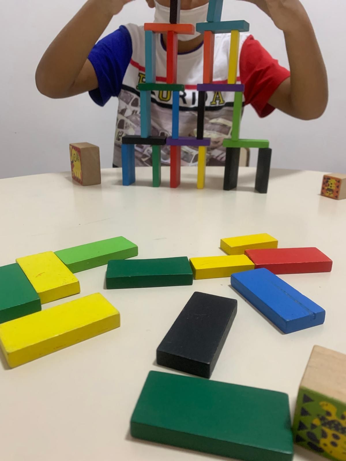 Niño construyendo una torre con bloques de madera de colores sobre una mesa, con bloques esparcidos alrededor.