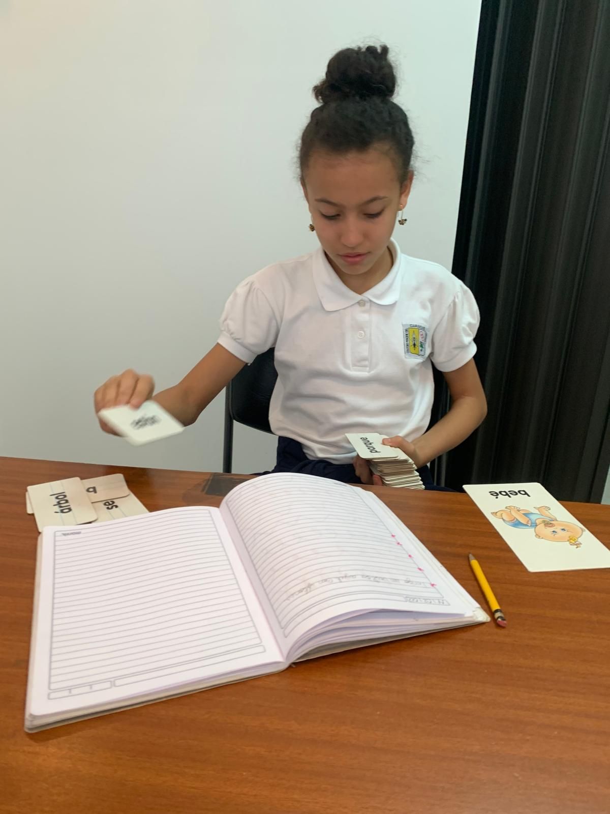 Chica sentada, estudiando cartas y un cuaderno en un escritorio. Camisa blanca y pelo recogido.