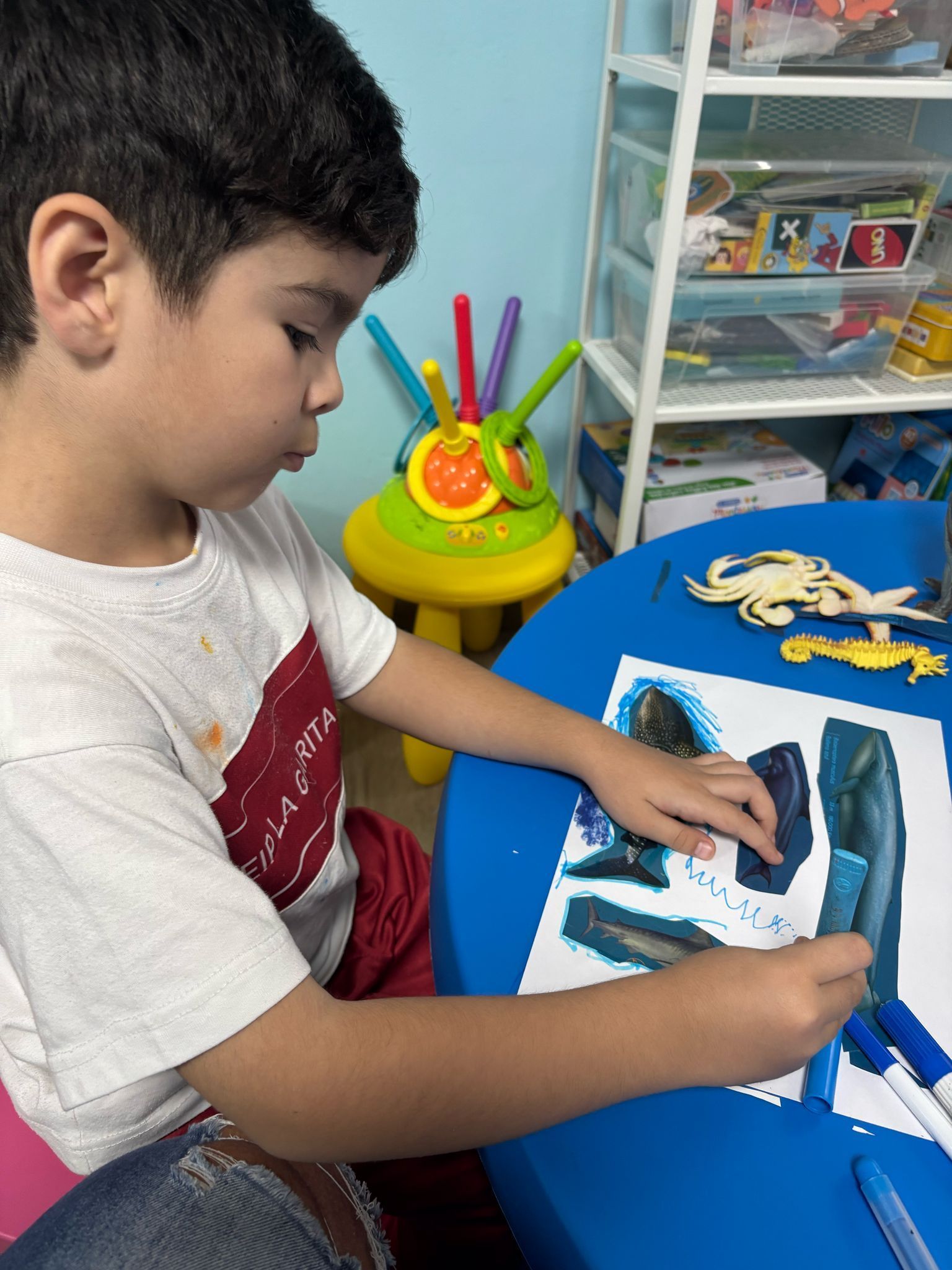 Niño dibujando en una mesa azul. Lleva una camiseta blanca y hay materiales de arte cerca.