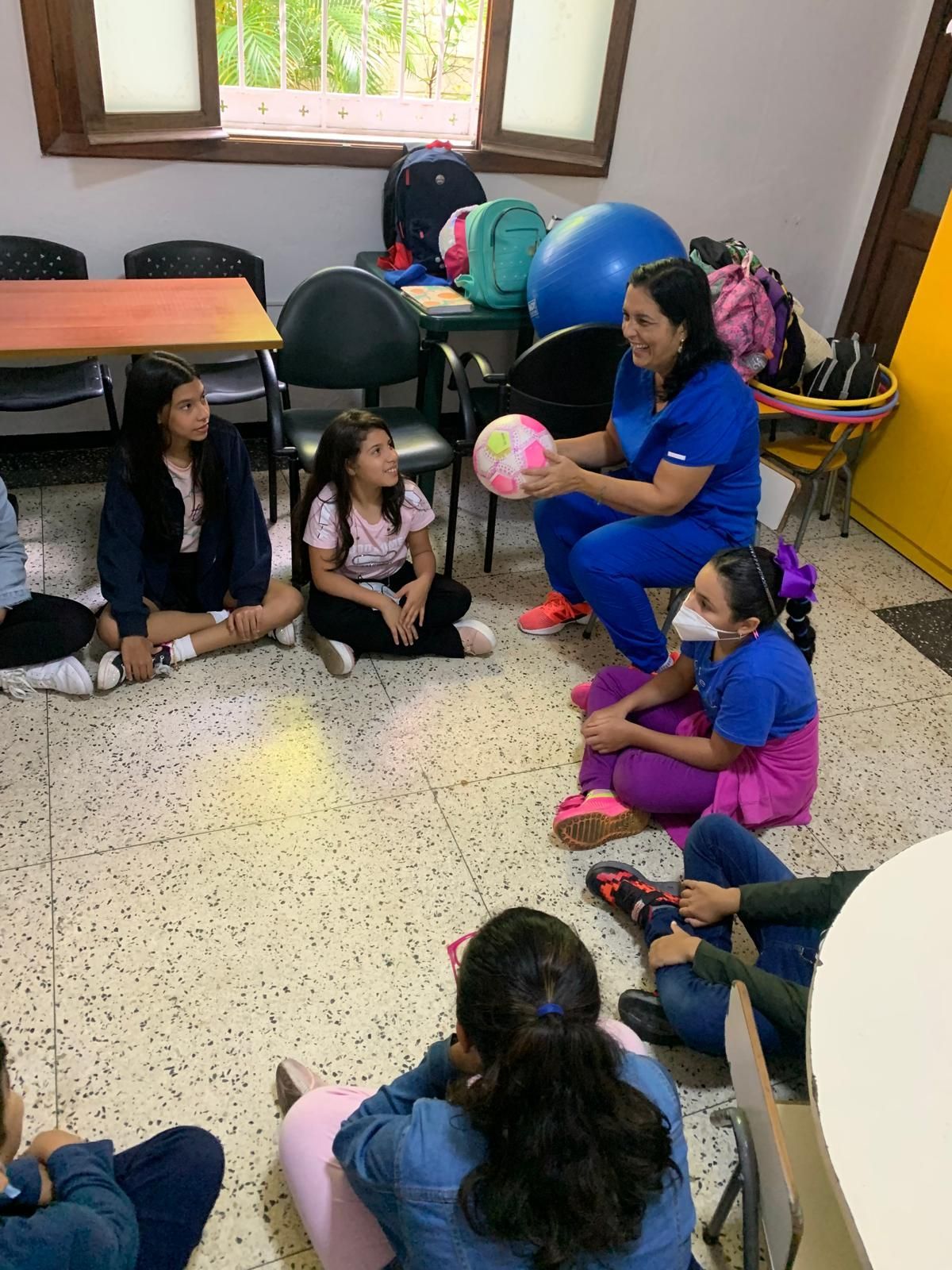 Una mujer con uniforme azul sostiene una pelota mientras habla con un grupo de niños sentados en el suelo.