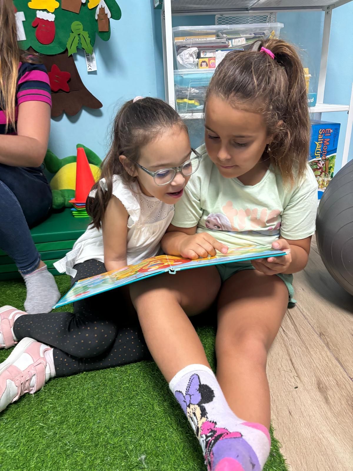 Dos chicas jóvenes leen un libro juntas, sentadas sobre una alfombra verde en el interior. Una lleva gafas.