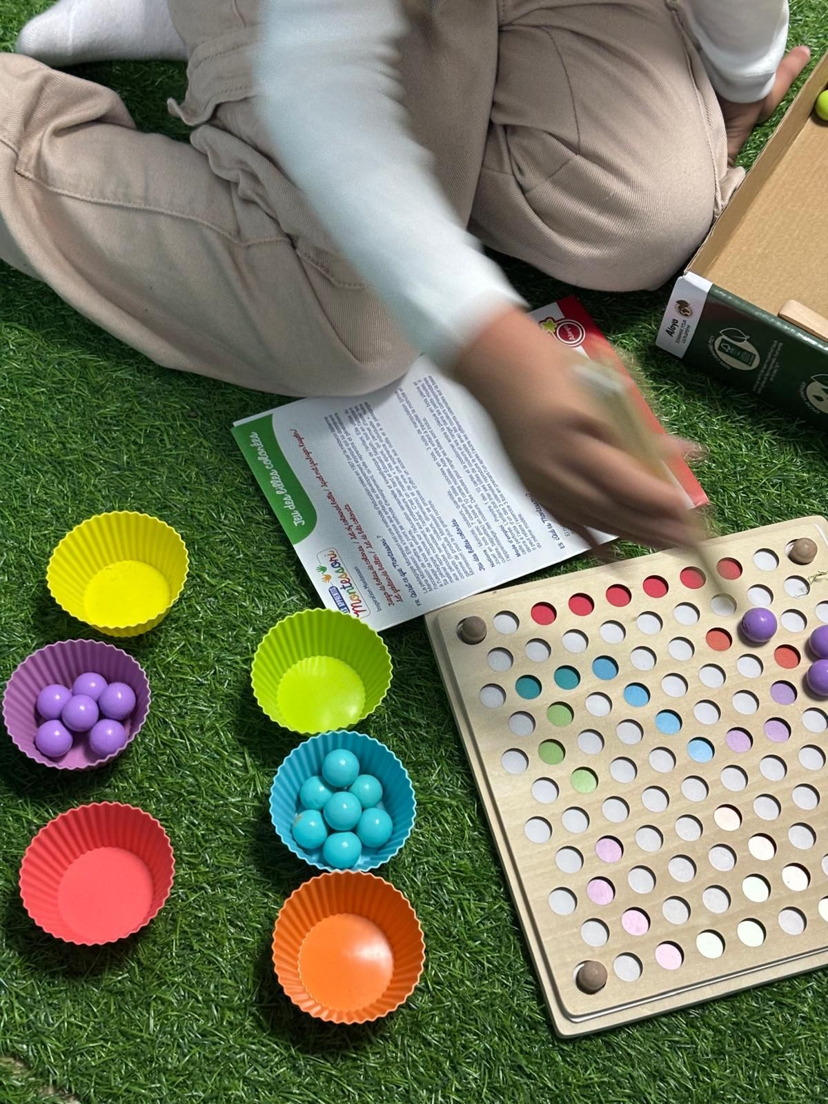 Un niño juega con una tabla de madera y pelotas de colores. Vasos brillantes sostienen las pelotas sobre una superficie verde.