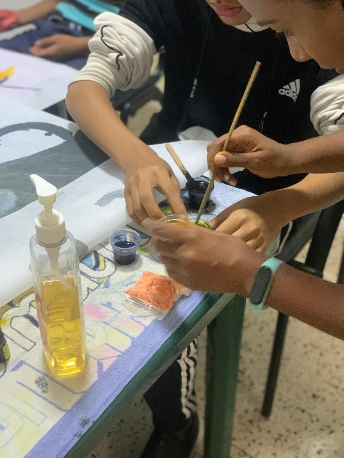 Dos personas pintan con pinceles y tinta. Hay una botella de jabón líquido sobre la mesa.