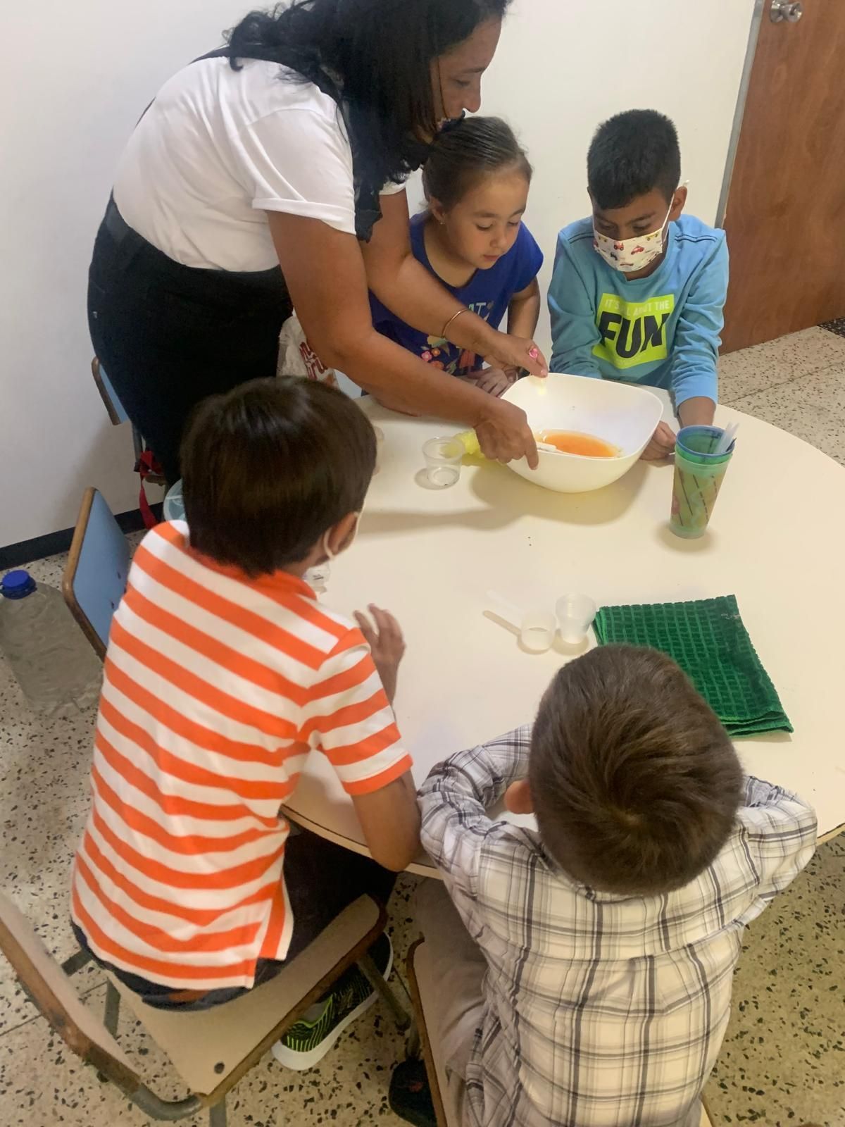 Maestra y niños mezclan ingredientes en una mesa. Los niños observan con curiosidad.