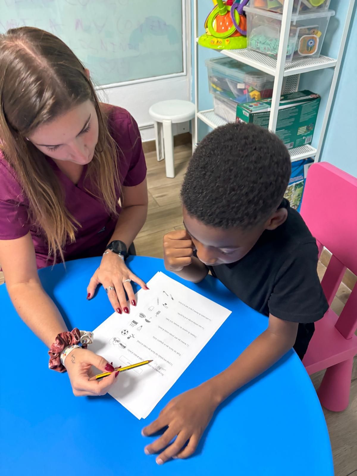 Mujer señala un papel con un lápiz, ayudando a un niño en una mesa azul.