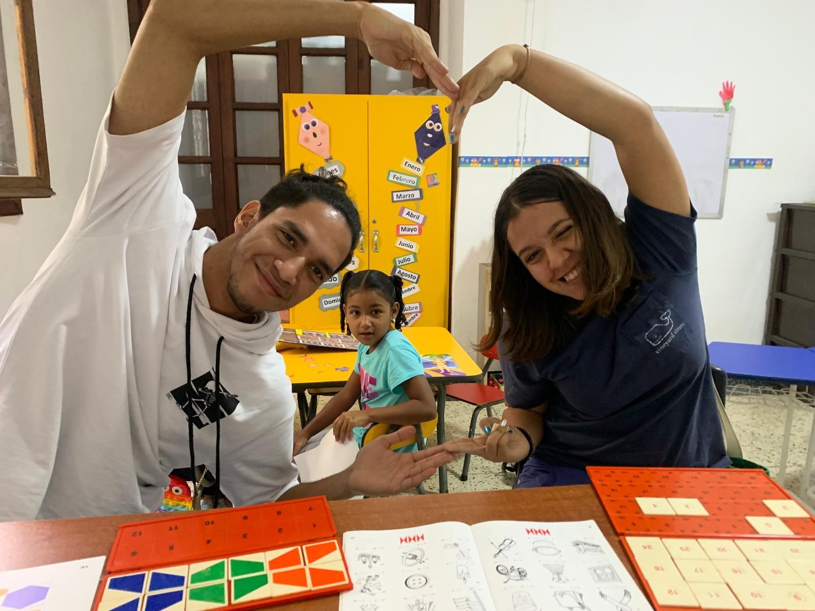Dos adultos y un niño sonriendo, formando un corazón con sus manos en un salón de clases.