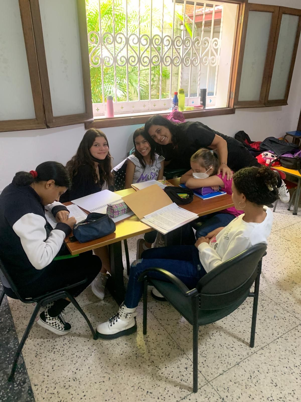 Grupo de niñas y un adulto en una mesa, estudiando junto a una ventana.