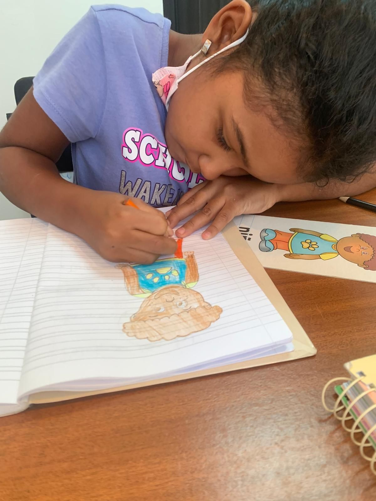Niña coloreando un cuaderno, con una máscara, sentada en una mesa.