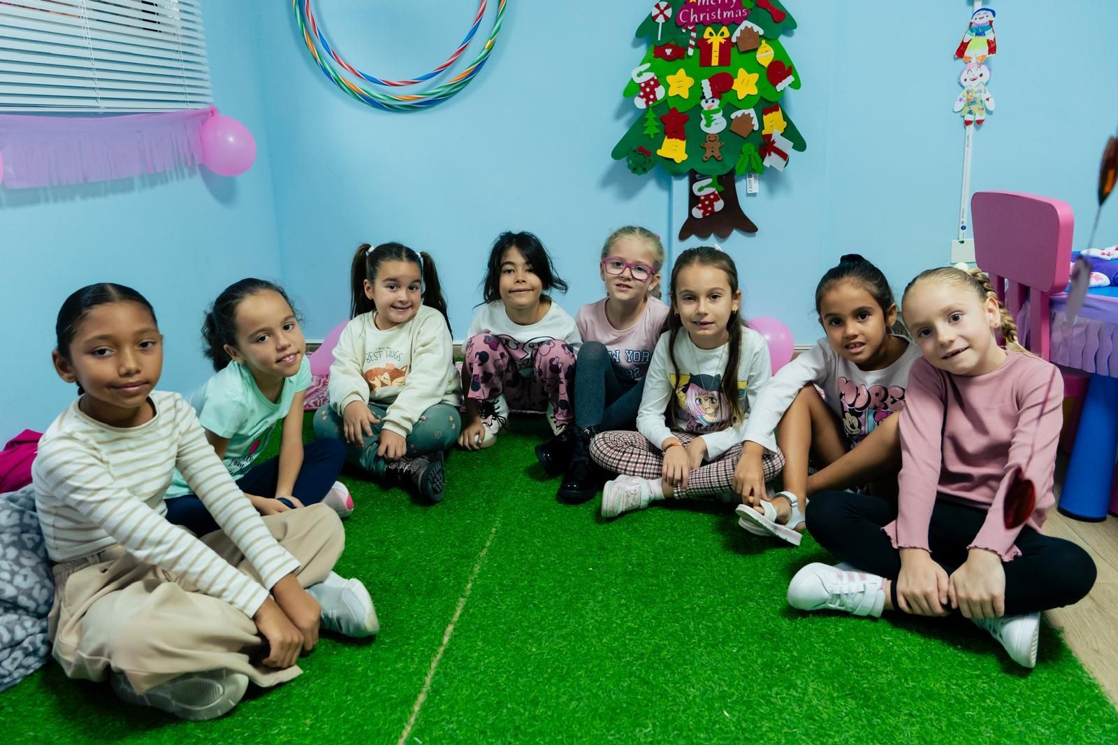 Un grupo de niñas sentadas sobre una alfombra verde en una sala de juegos con decoraciones navideñas.