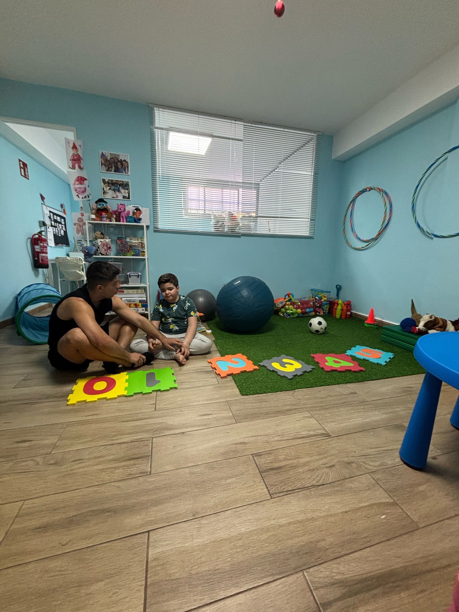 Hombre y niño sentados en el suelo de la sala de juegos. Juguetes coloridos, paredes azules y césped artificial.