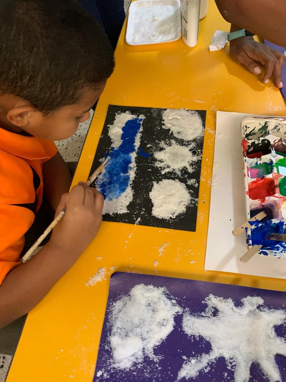 Niño pintando azul sobre papel negro con sal, en una mesa amarilla. Pinturas y materiales cerca.