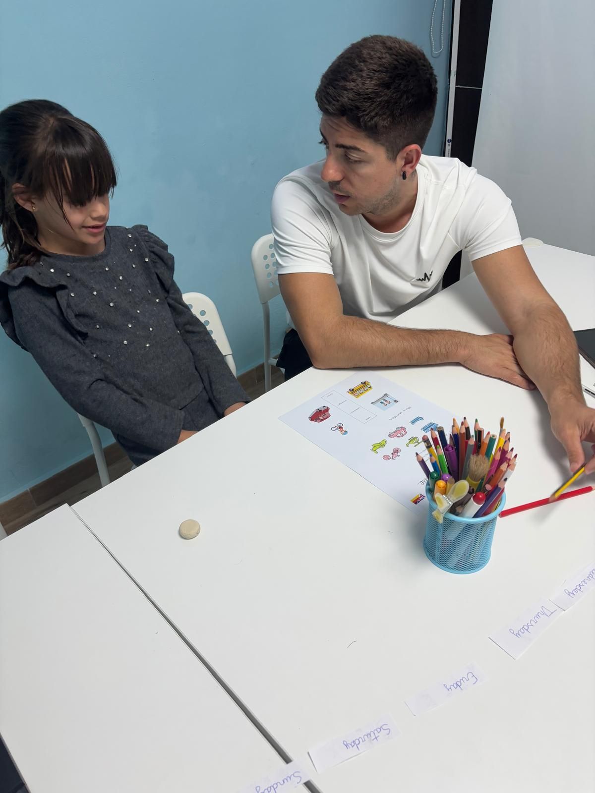 Un joven y una niña están sentados en una mesa con lápices y un dibujo; están mirando el papel.