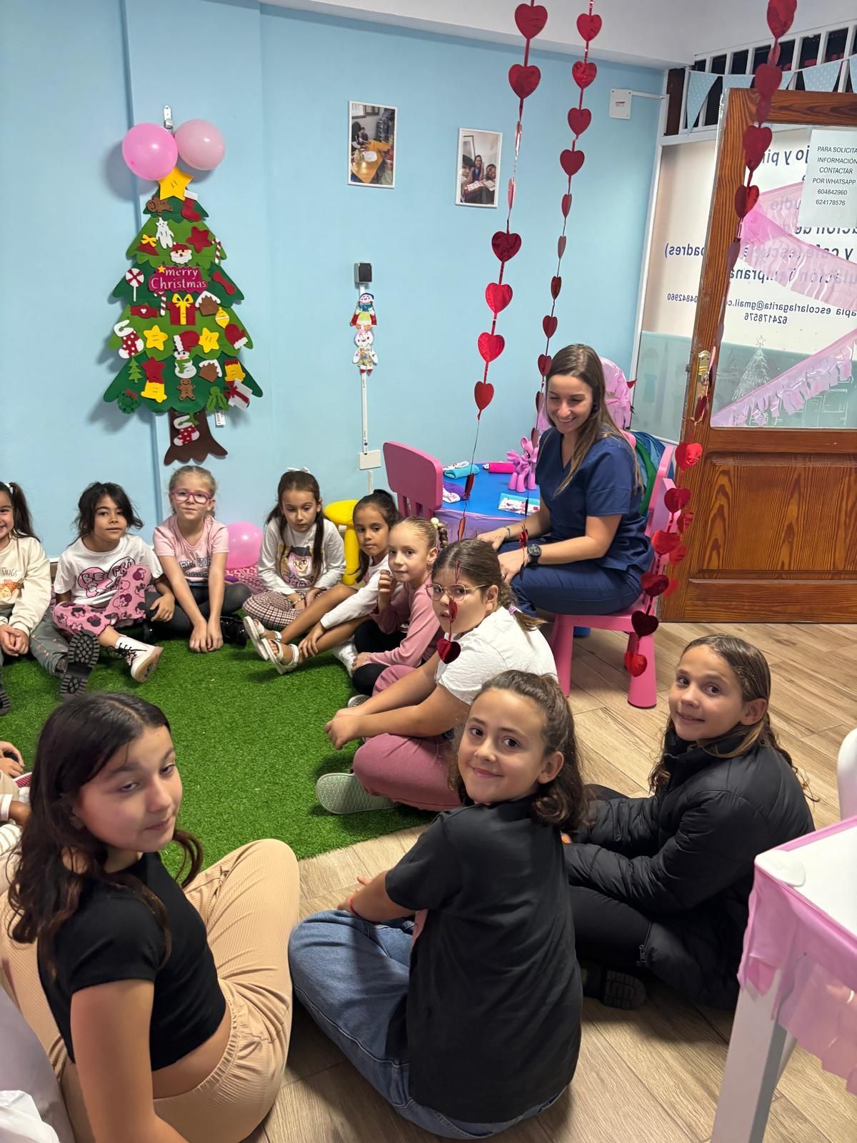 Unos niños y una mujer están sentados sobre una alfombra verde. Están en una habitación decorada para una fiesta, posiblemente una celebración de San Valentín.