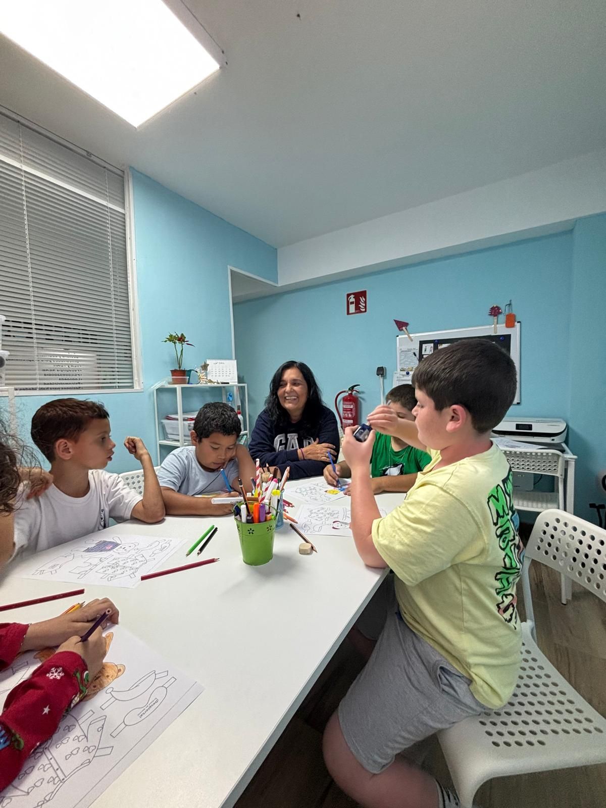 Niños y un adulto en una mesa, coloreando y dibujando. Un niño toma una foto. Paredes azul claro, habitación luminosa.