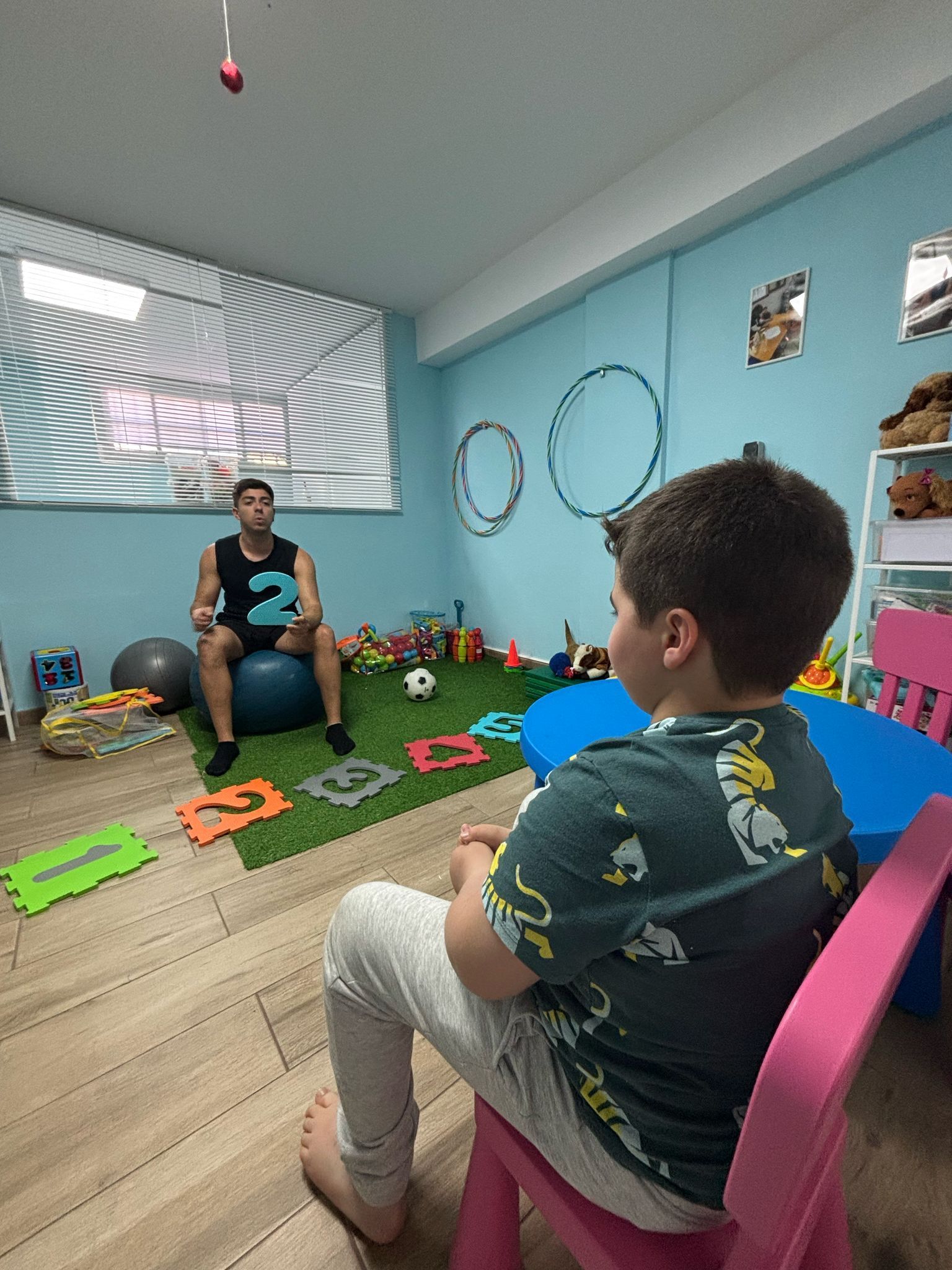 Un niño observa a un hombre que sostiene un número 2 azul, sala de terapia interior.