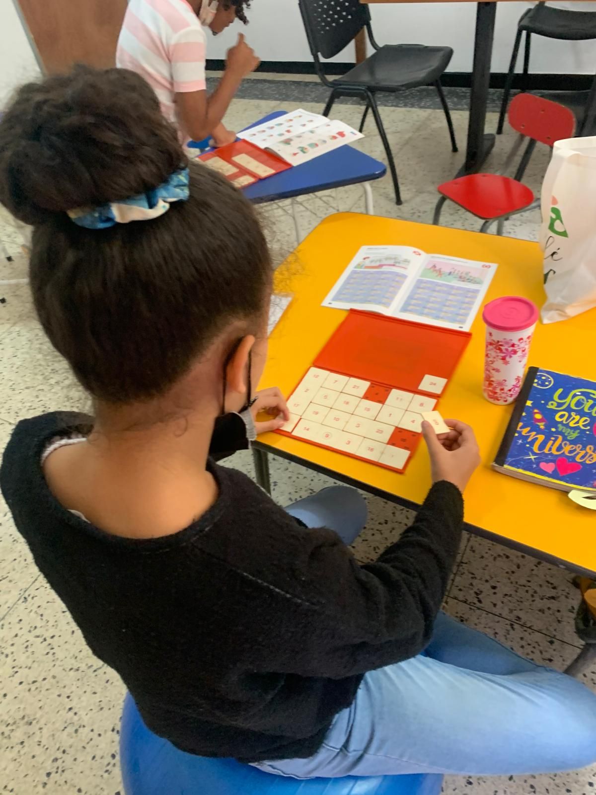 Chica en una mesa amarilla, con mascarilla, armando un rompecabezas. Libro y taza cerca.