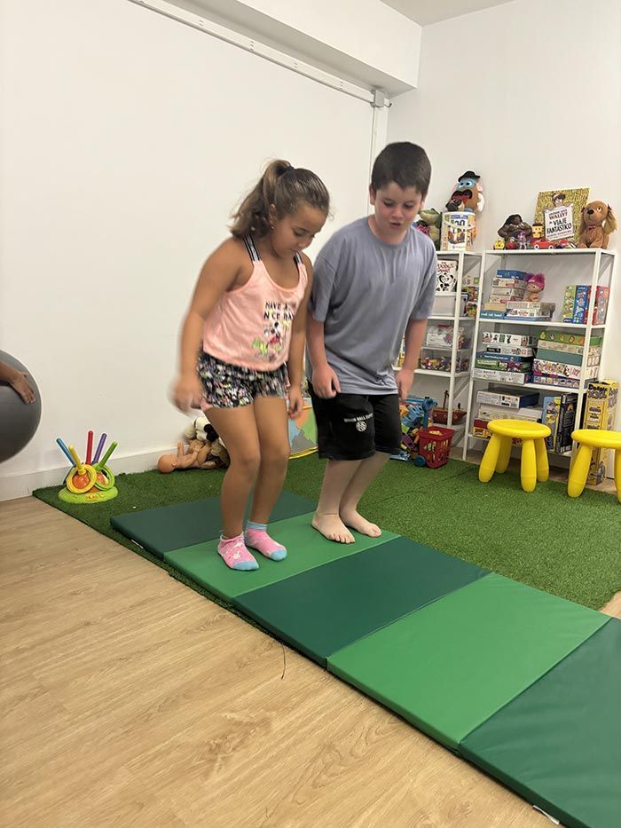 Dos niños saltan sobre una colchoneta verde en el interior. La niña lleva pantalones cortos y una camiseta sin mangas, el niño lleva pantalones cortos y una camiseta gris.