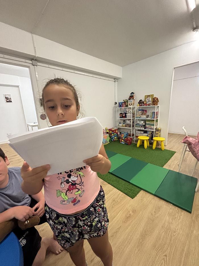 Una niña lee un papel, posiblemente en un aula. Se ven otros niños, tapetes y juguetes.