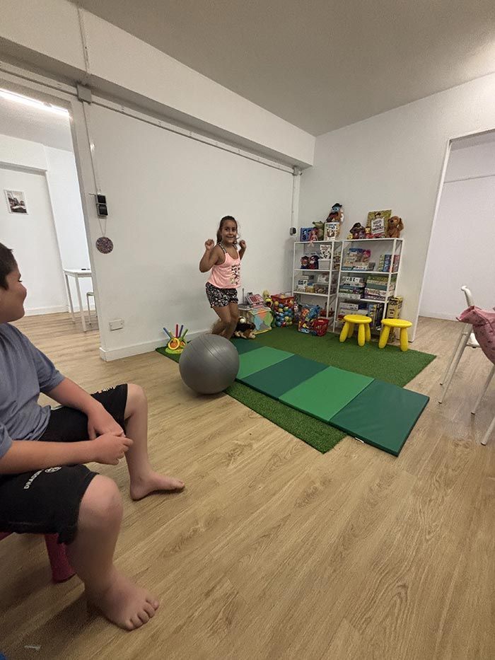 Un niño hace ejercicio en una habitación, mientras otro niño observa. Se ven juguetes y equipos.