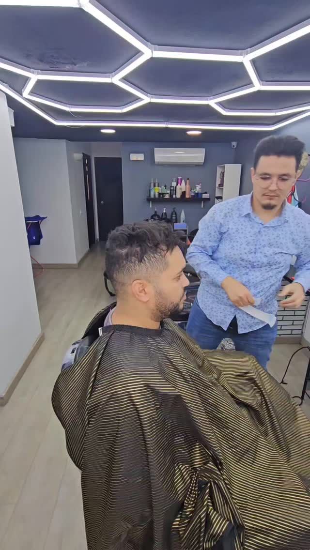 Un barbero corta el pelo a una clienta en un salón. La clienta está sentada, con una capa. El barbero lleva una camisa azul.