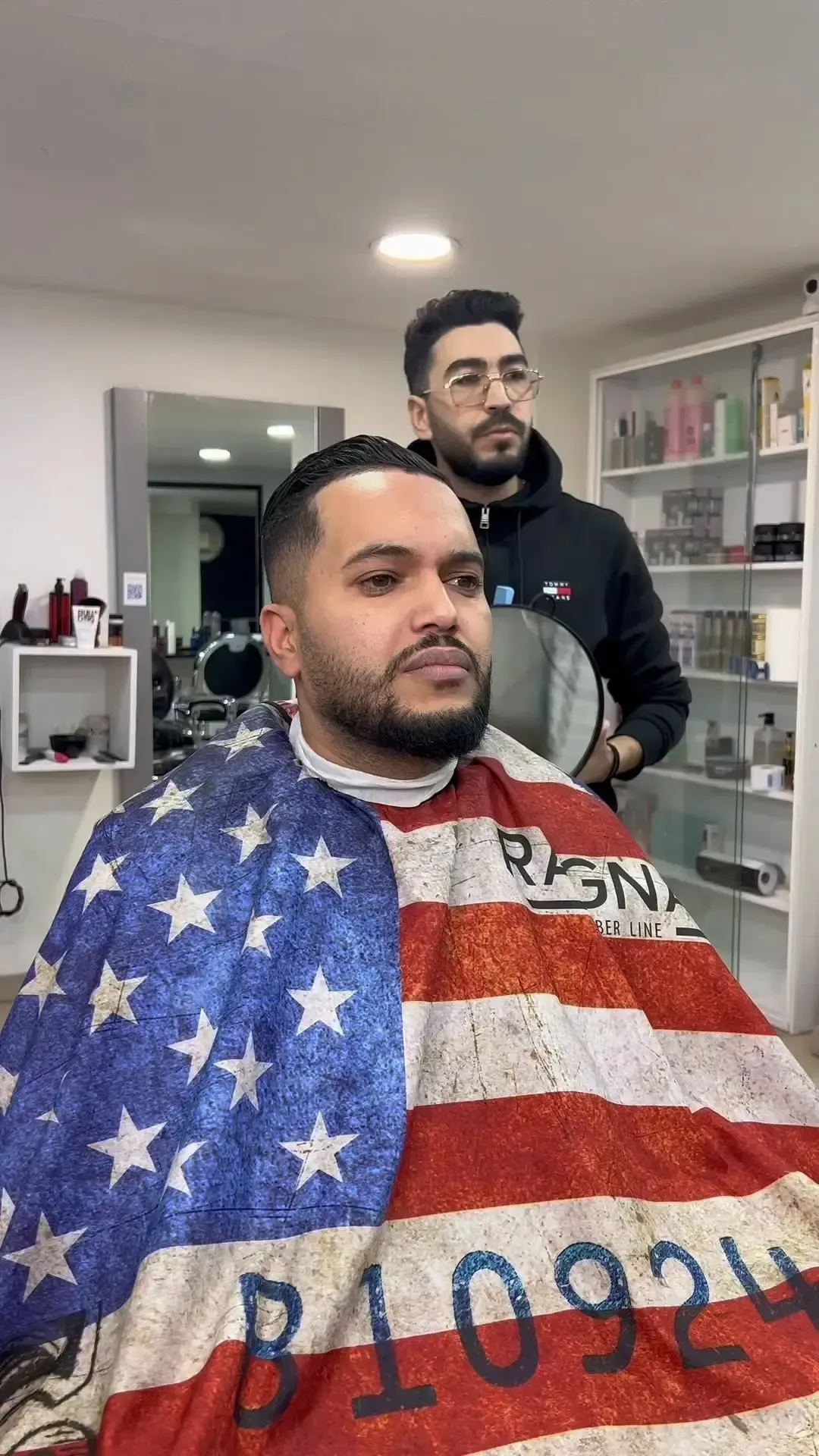 Hombre en silla de barbero con capa con bandera estadounidense; barbero sosteniendo un espejo.