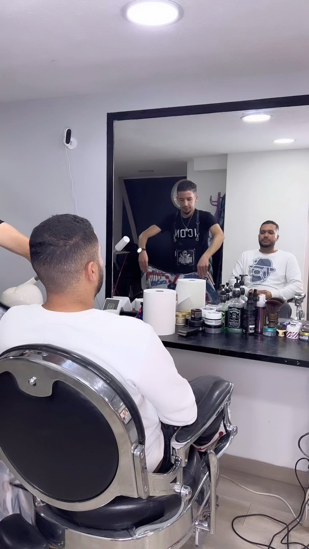 Un barbero corta el pelo en una peluquería. Un hombre sentado en una silla se mira al espejo. Otro hombre observa.