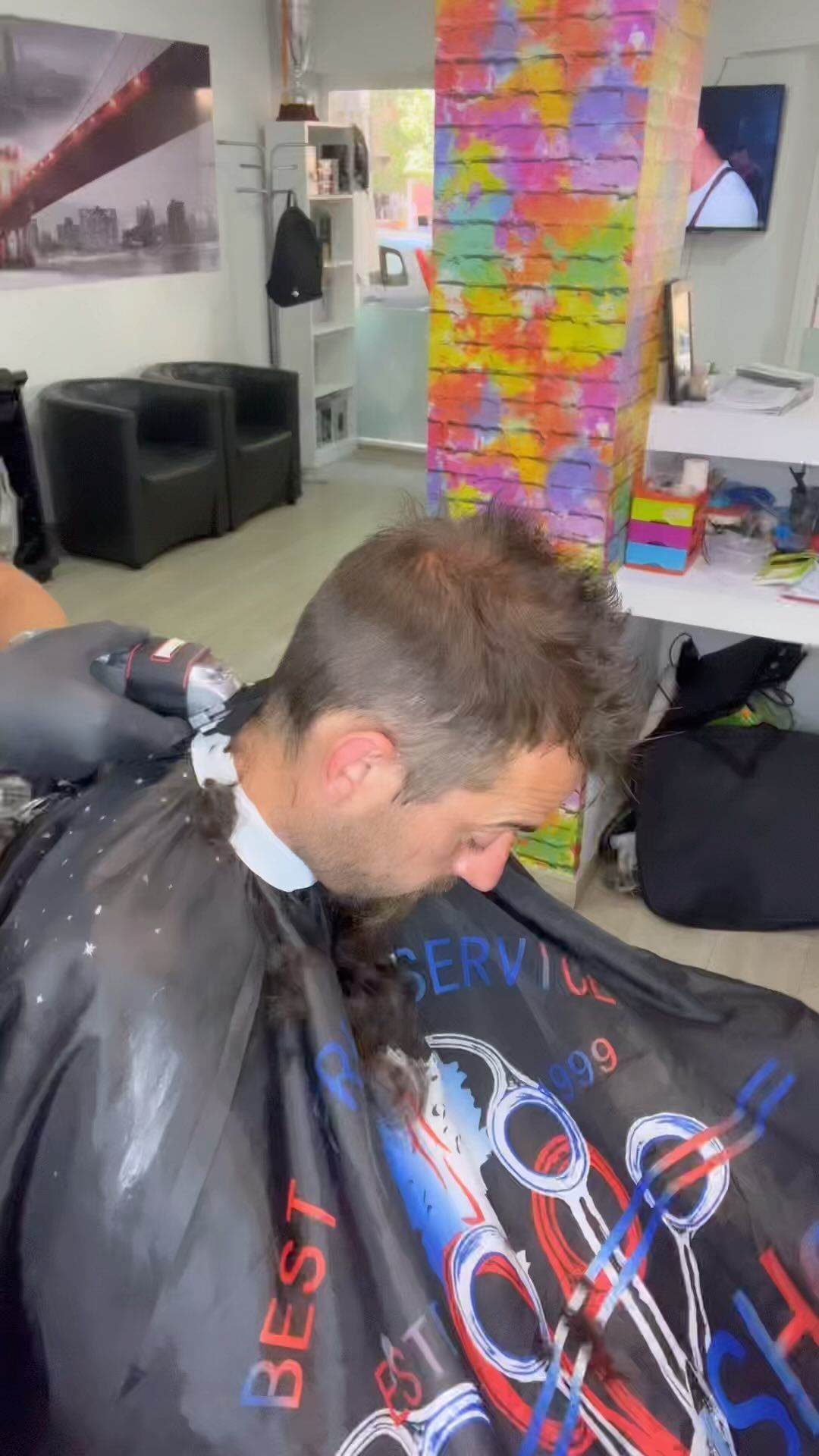 Un hombre se corta el pelo en una barbería. Lleva una capa y una maquinilla en uso. Al fondo, una pared colorida.