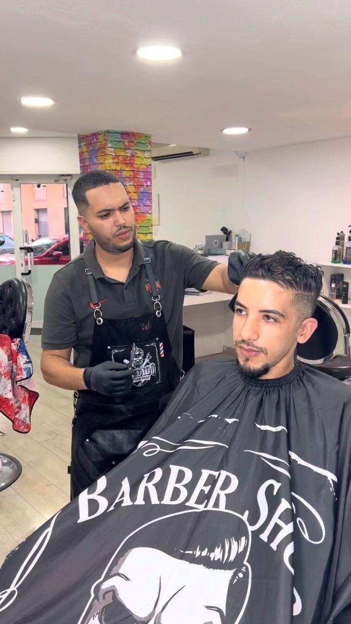 Un barbero corta el pelo a un cliente en una peluquería. El cliente lleva una capa de barbero y mira al frente.