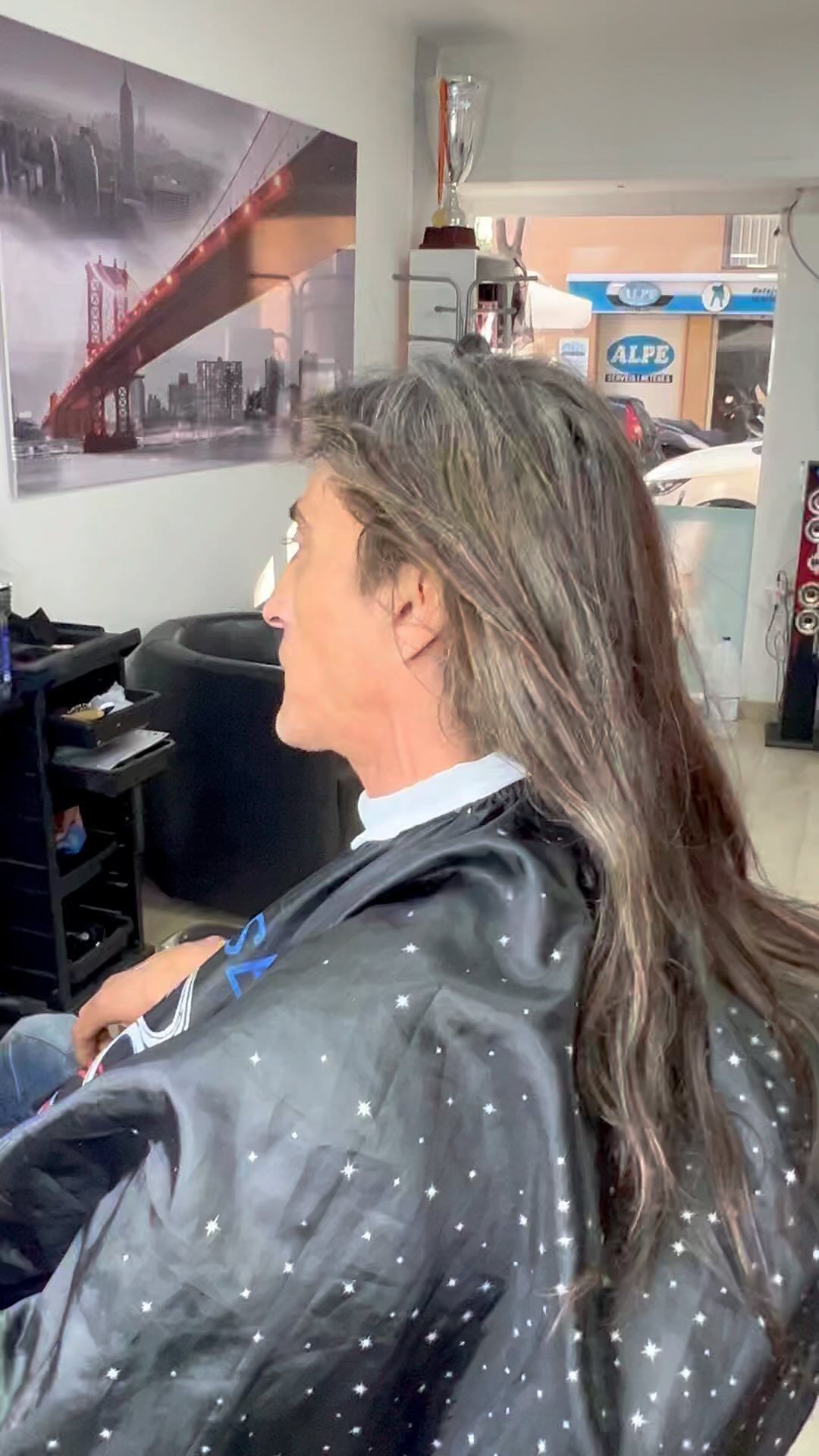 Persona con cabello largo y gris en una silla de salón, vistiendo una capa negra con tachuelas plateadas.