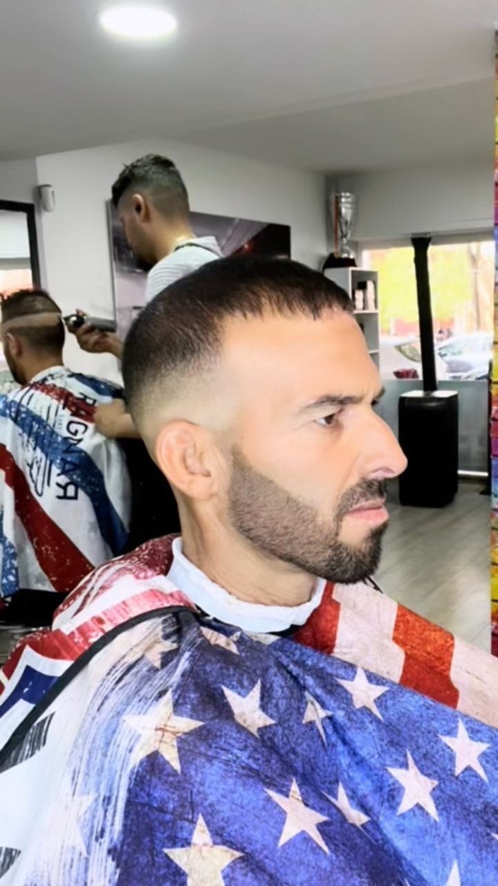 Un hombre se corta el pelo en una barbería; lleva una capa con la bandera estadounidense. Otra persona le está cortando el pelo.